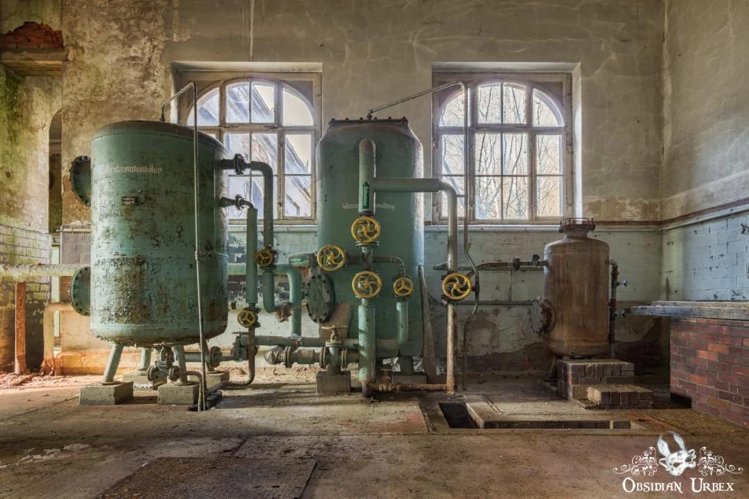 An abandoned textile factory turbine hall features two large teal tanks with yellow valves and pipes, alongside a smaller brown tank.