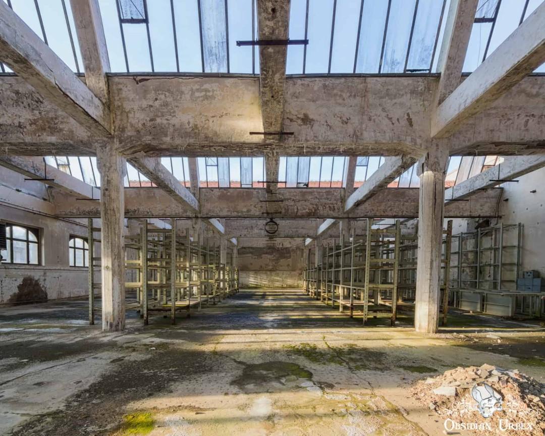 A wide-angle view of an abandoned industrial hall with concrete pillars, glass roof, and rows of empty metal shelving.