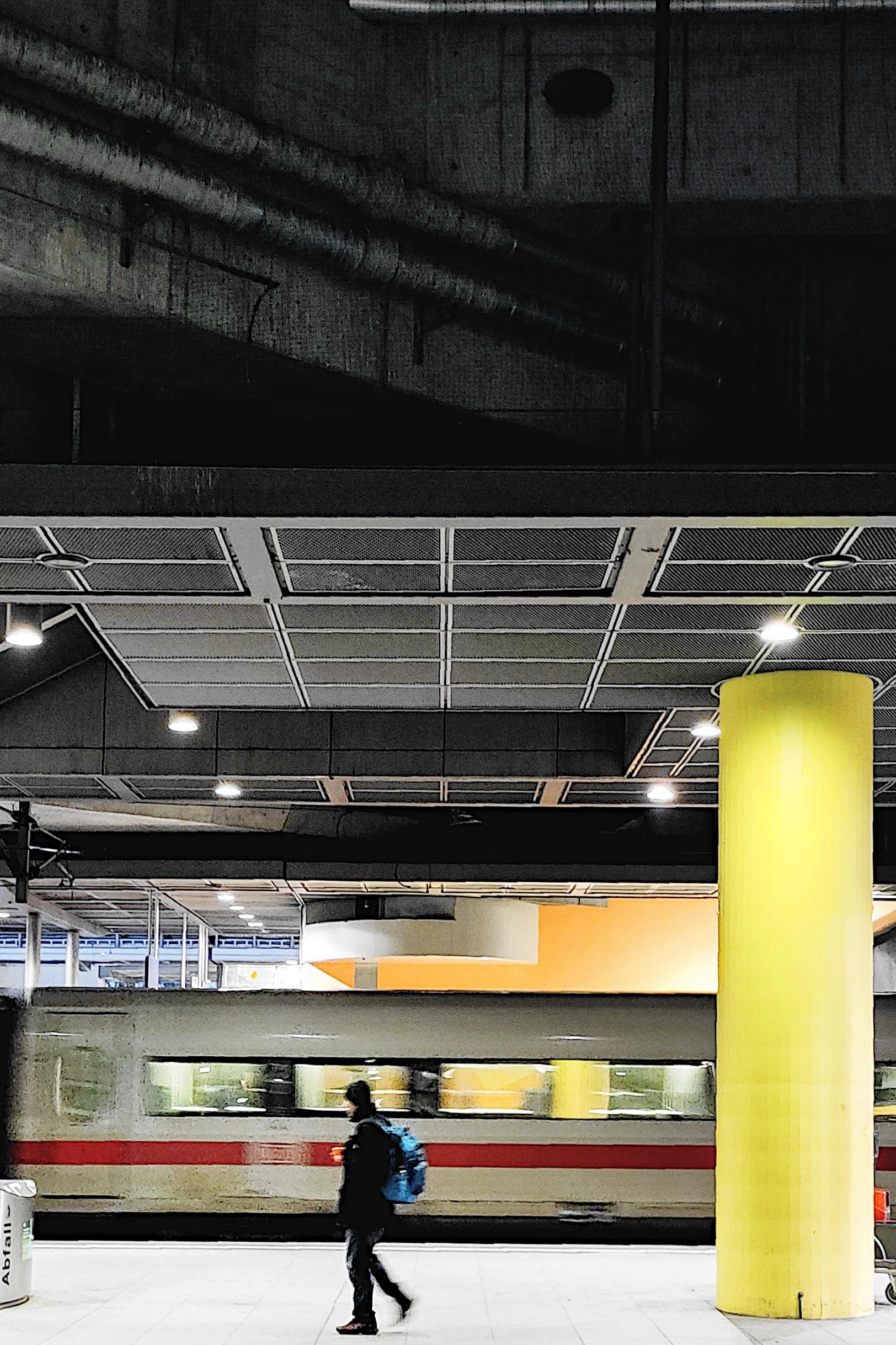 The interior of a covered railway station or underground platform with a dark concrete ceiling, exposed ducts and grid panels lit by round ceiling lamps. A person carrying a blue and black backpack walks across the pale platform floor in the lower centre, captured with slight motion blur. Behind them a train with a red stripe stands or moves along the track, also blurred. A tall bright yellow cylindrical column dominates the right side of the frame. A waste bin labelled "Abfall" stands at the lower left. The scene is mostly dark with the yellow column and platform floor standing out.