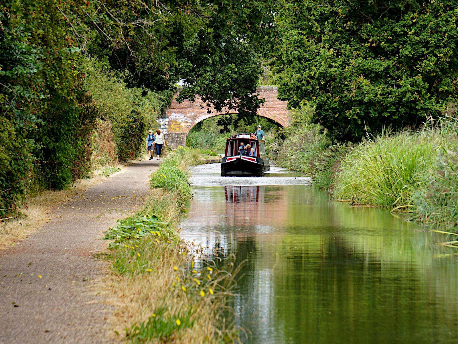 A boat on a canal. it has just passed under a brick bridge and passing under some overhanging trees. There are 2 people in the front of the boat, a lone person at the back on the tiller.
2 people are walking beside it on the tow path. The boat and vegetation on the opposite side of the canal are reflected in the water.
