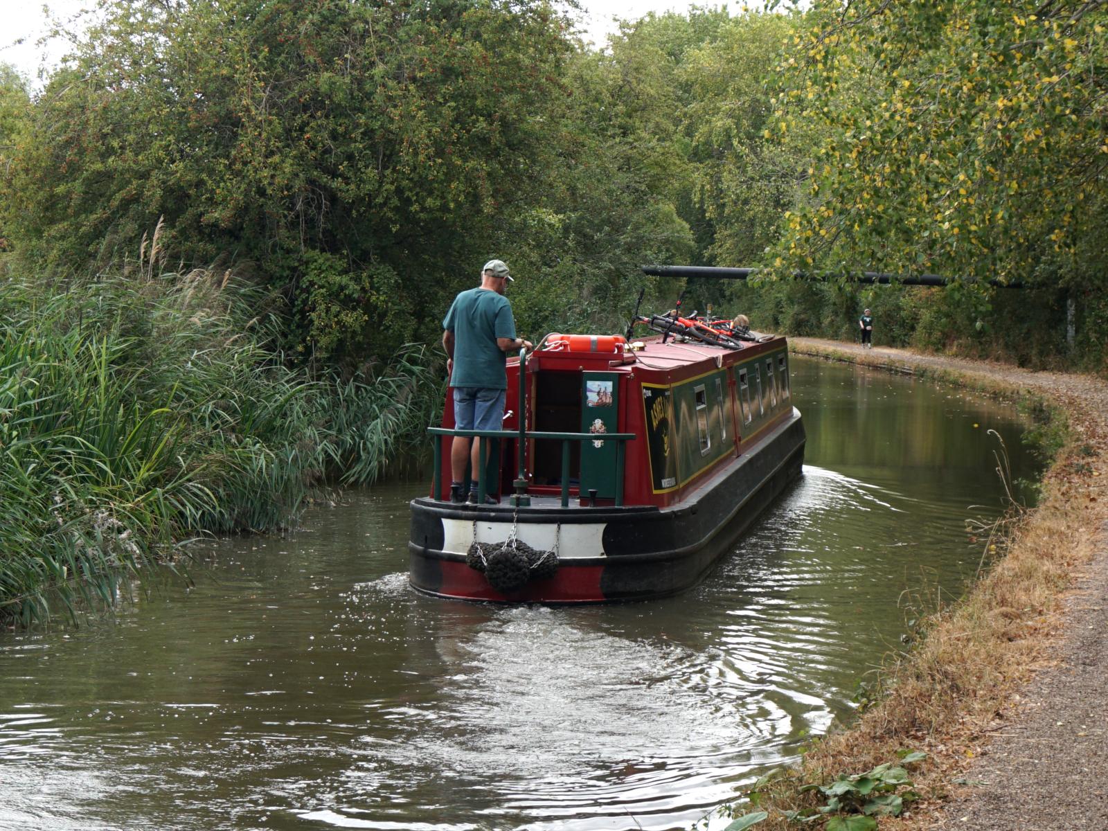 A view of the back of a canal boat moving away from the camera, there is a man who is steering it standing at the back of the boat.
The canal and tow path veer away to the left underneath a metal pipe suspended above the canal. (I believe it's a gas pipeline)
There are rushes and trees on the left of the canal and trees beyond the tow path
