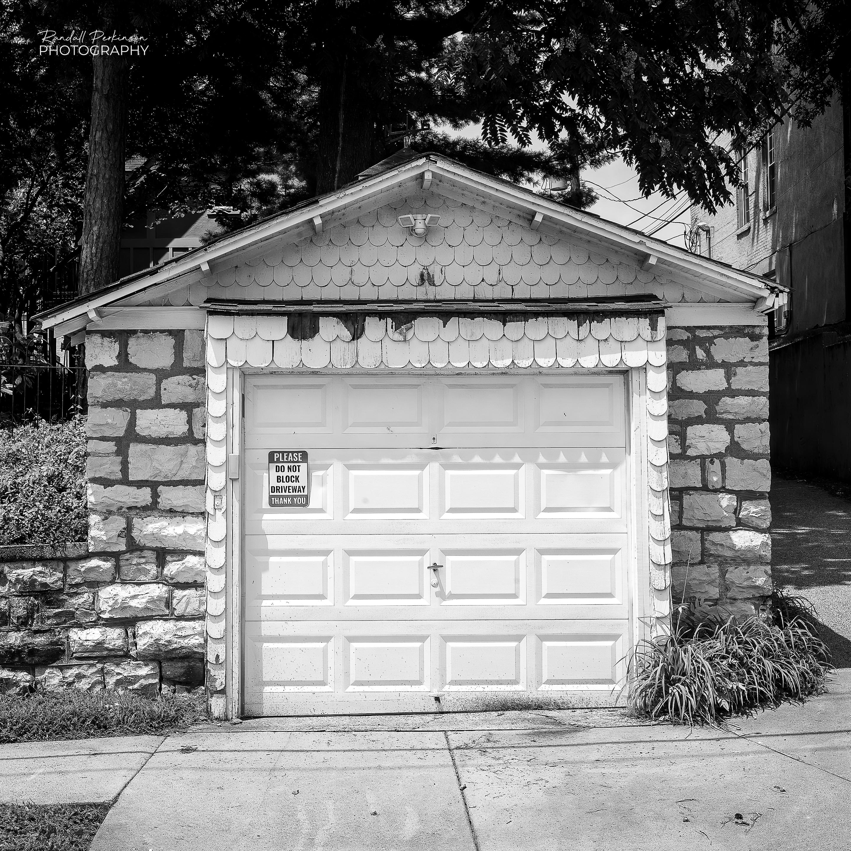A black and white view of a one car garage made of stone with white wood shingles covering the upper front.
