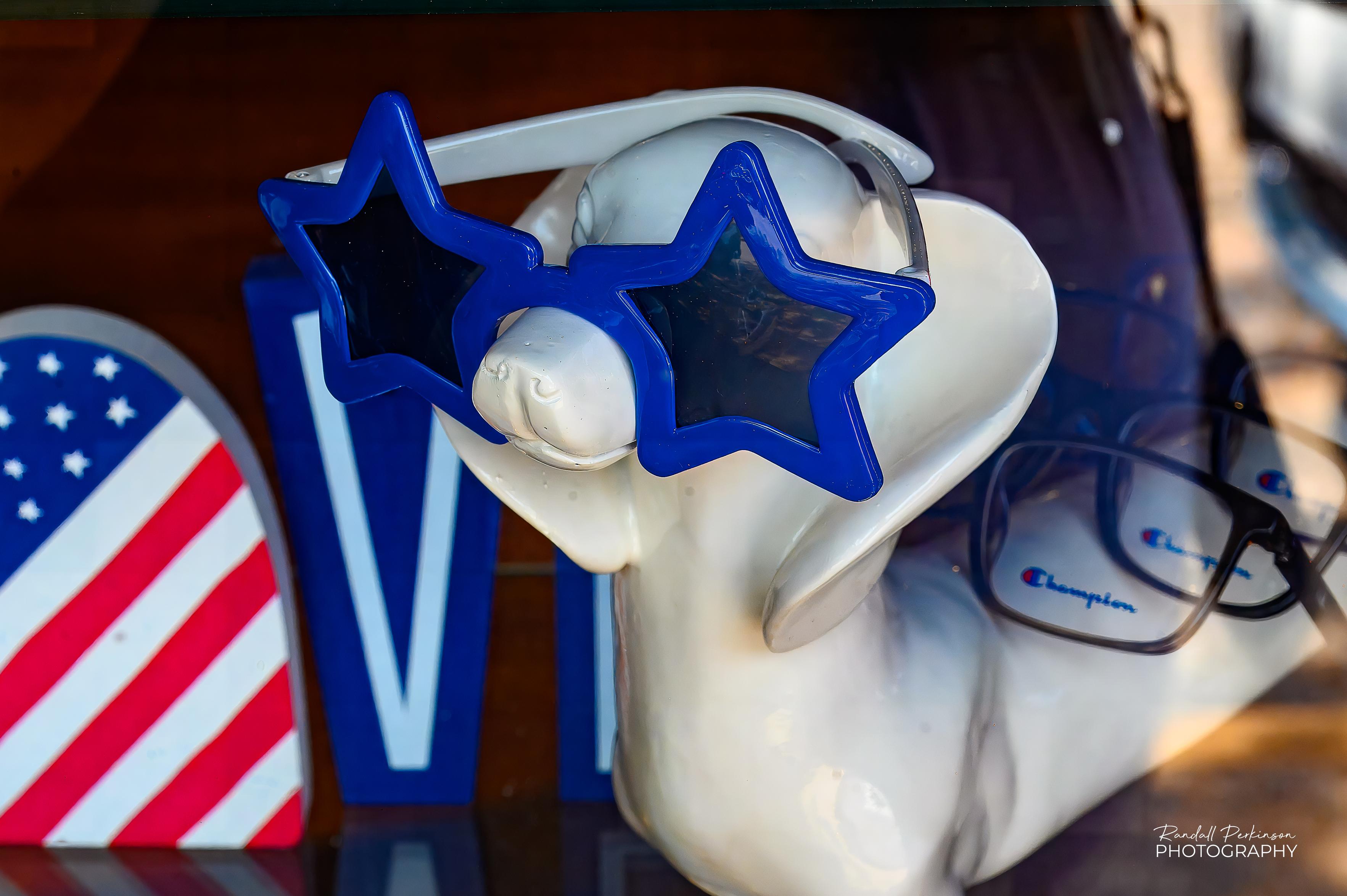 An eyewear shop window display with a ceramic dachshund wearing blue star sunglasses with other glasses laying across it's back.