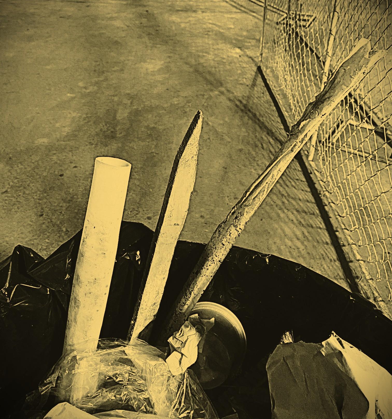 Monochrome image of a wooden stake and other discarded materials in a trash can in the second sublevel deck of an underground parking structure in Akron, Summit County, Ohio, USA. Image shows the top arc of the trash can and a plastic pipe, a fiberglass marker, and a wooden stake protruding above the rim, the concrete floor of the deck beyond and a chain link fence to the right.