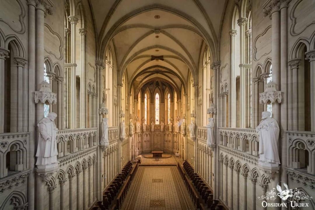 An ornate, vaulted hall, possibly an abandoned church, stretches towards sunlit windows. White statues line decorative balustrades overlooking empty benches.