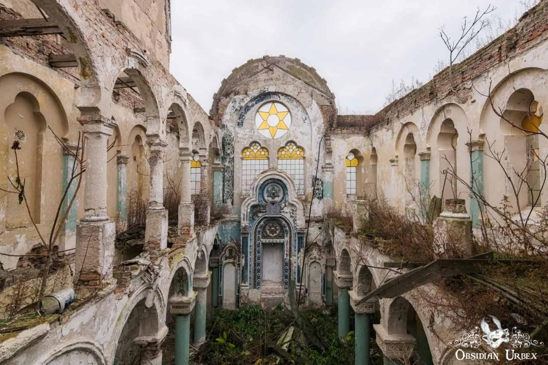 An abandoned synagogue interior features a Star of David stained-glass window. Arches and pillars line the crumbling hall, with vegetation growing throughout