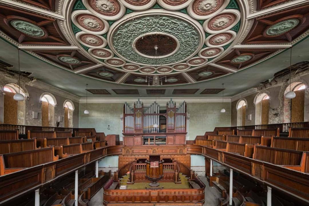 An abandoned, glorious chapel interior features a grand ornate ceiling, rows of wooden pews, and a large pipe organ