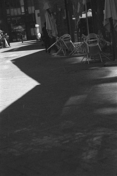 Ilford HP5 Plus 400 (FF)

English
A black and white  photograph of  street scene. Several tables and chairs can be seen stacked upon each other outside of a shop. Pedestrians can be seen in the background. The contrast of light and shadow on the ground is strong.

中文
一张街景的黑白照片.照片中的桌椅相互堆叠在一家店的门口.背景中行人隐约可见.地面的光影对比强烈.