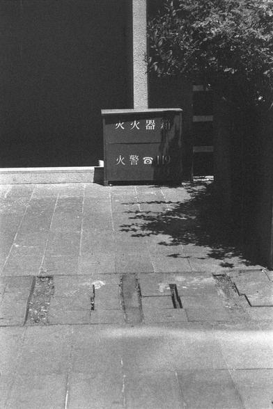 Ilford HP5 Plus 400 (FF)

English
A black and white photograph of a street scene. A fire hydrant storage stands in front of the door/wall. Dense foliage can be seen on the right side of the photo. The ground is  layered with grid tiles.

中文
一张街景黑白照片.墙边/床边可以看见灭火器的工具箱.照片右侧有大量的植被.地面是格状的地砖.
