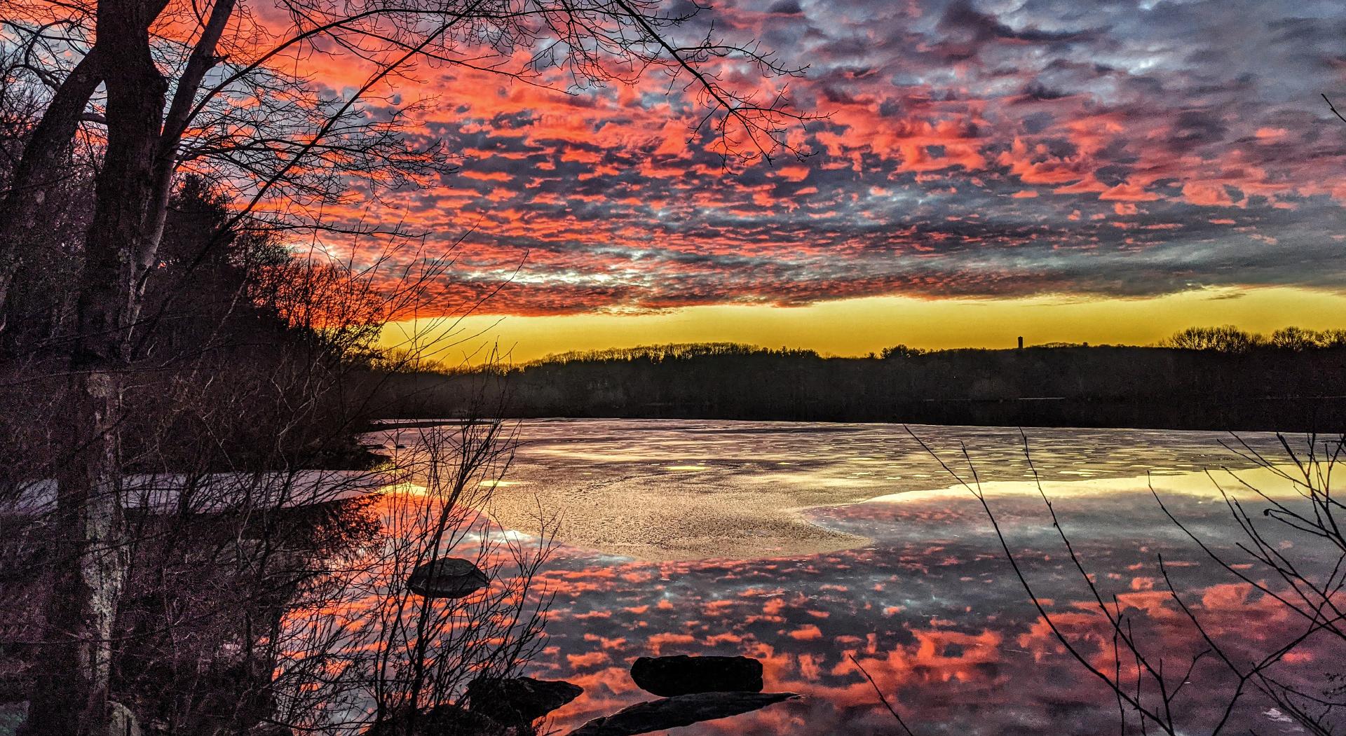 Banks of gray clouds are retreating eastward, leaving a swath of clear sky along the western horizon. The recently set sun is tinting that clear sky pale gold, while tinting the bottoms of many of the clouds a bright pink. We are standing beside a big lake, on a wooded rocky shoreline. A think skim of ice with dozens of melted patches covers much of the lake, a wide swath near our shore is completely thawed, mirroring the colorful sky. A forest of leafless hardwoods with a few evergreens encircles the lake.