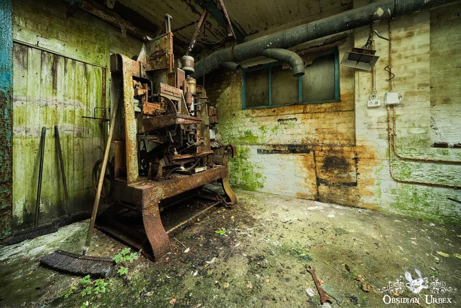 rusty machine in a room with decay, the walls are green with algae. there is an old broom leaning on the machine