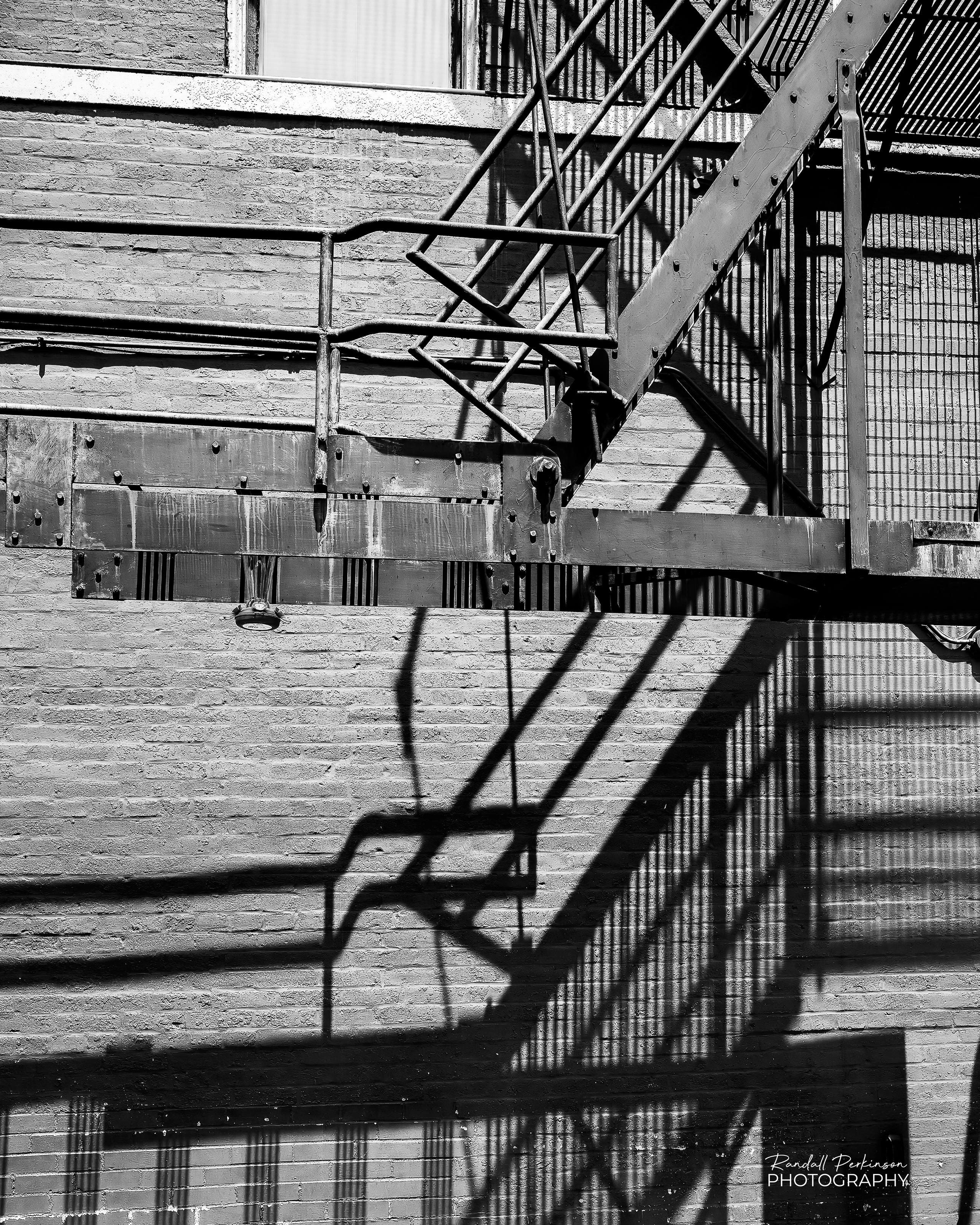 A black and white view of metal fire escape stairs on a brick building.  It's throwing a detailed shadow against the side of the building.