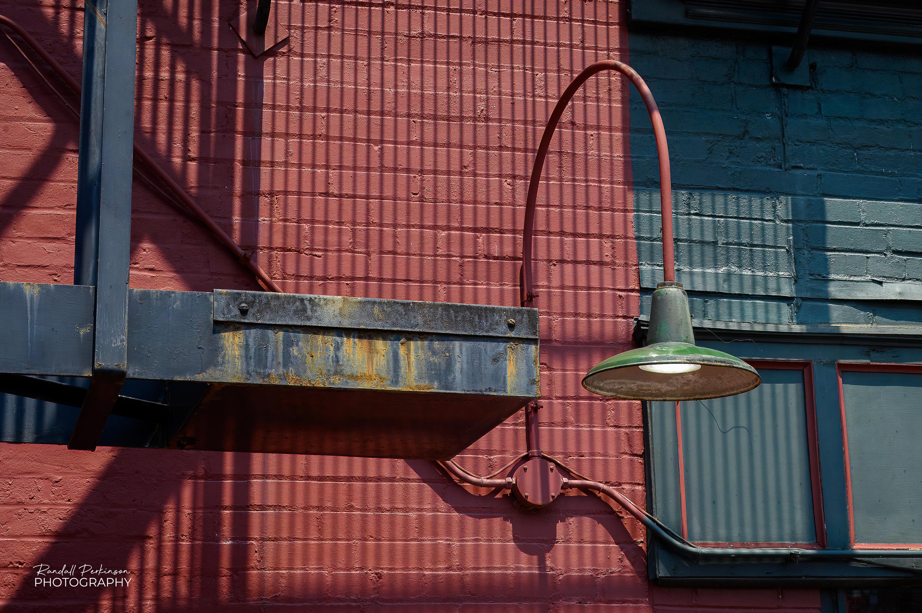 View of the outside of a red brick building with a portion of a metal fire escape on the left, a light hanging from a curved piece of pipe and some windows with some blacked-out bricks above it.