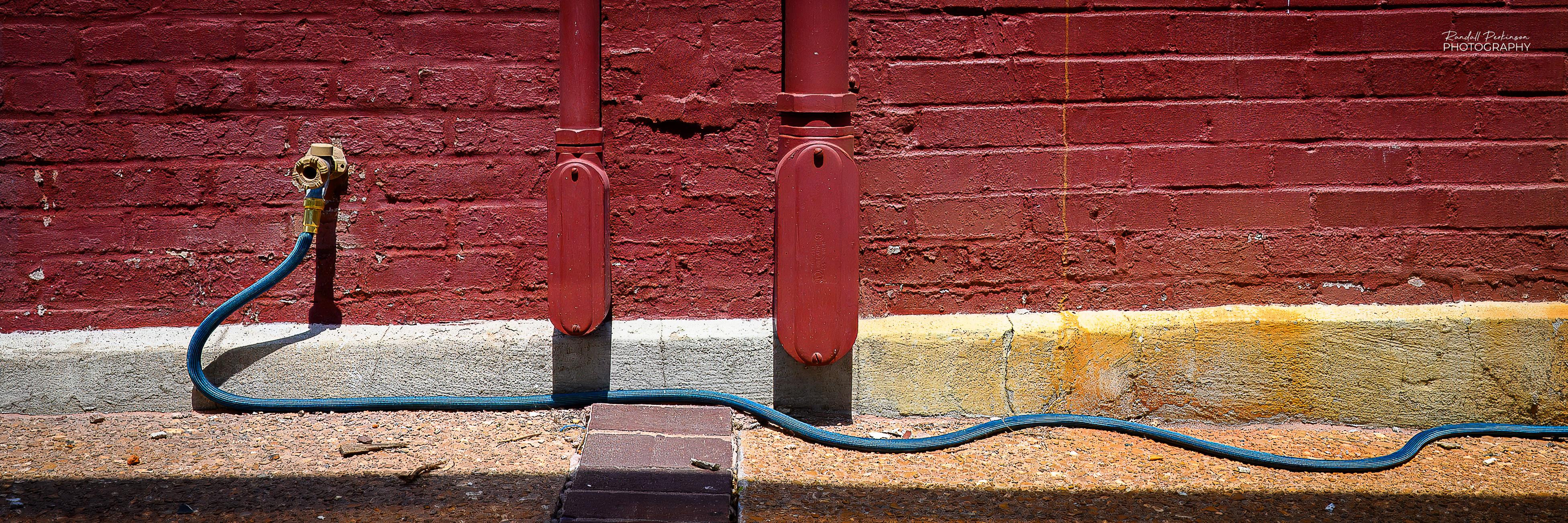 A very wide image of the bottom of a brick wall painted red.  In the center are two electrical conduits coming down from above with a small box where they enter the building.  To the left is an outdoor faucet with a green hose than drops down to the sidewalk and snakes along the bottom edge of the building to the right.