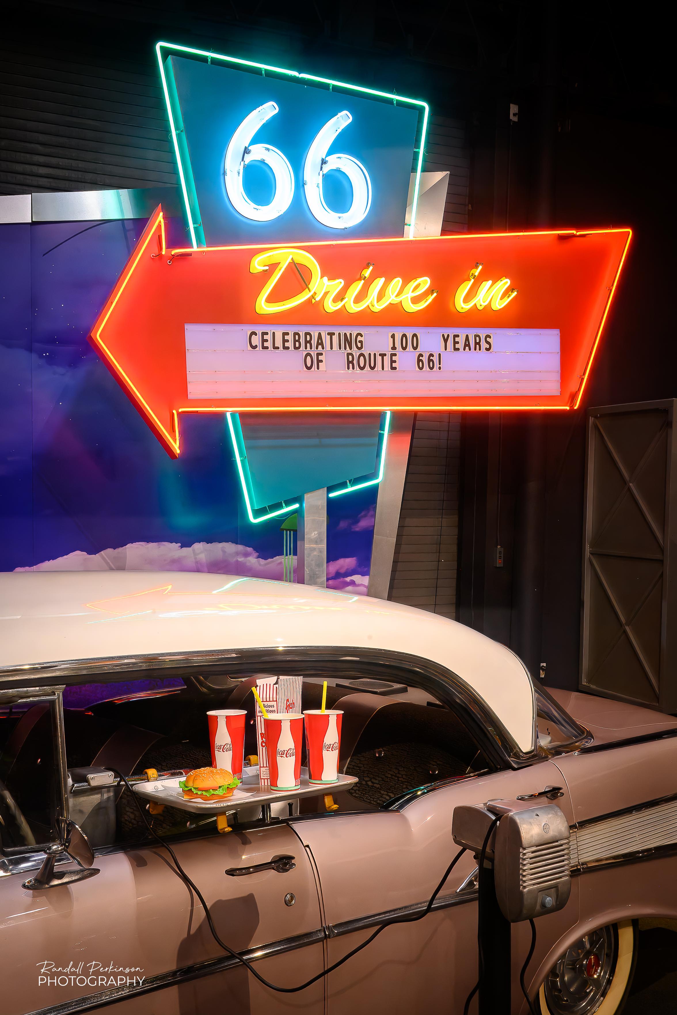 An exhibit at The National Museum of Transportation in recognition of the 100th Anniversary of Route 66.  It has a "66 Drive in" neon sign in the background and a mid-50's Cadillac with a window tray holding a hamburger and drinks.