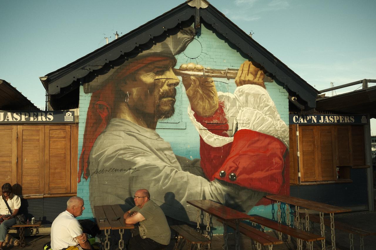 A picture of a resturant called Cap'n Jaspers. On the side of the building has a painting of a captain looking down an spyglass.
There are some people sat on benches in the foreground.
