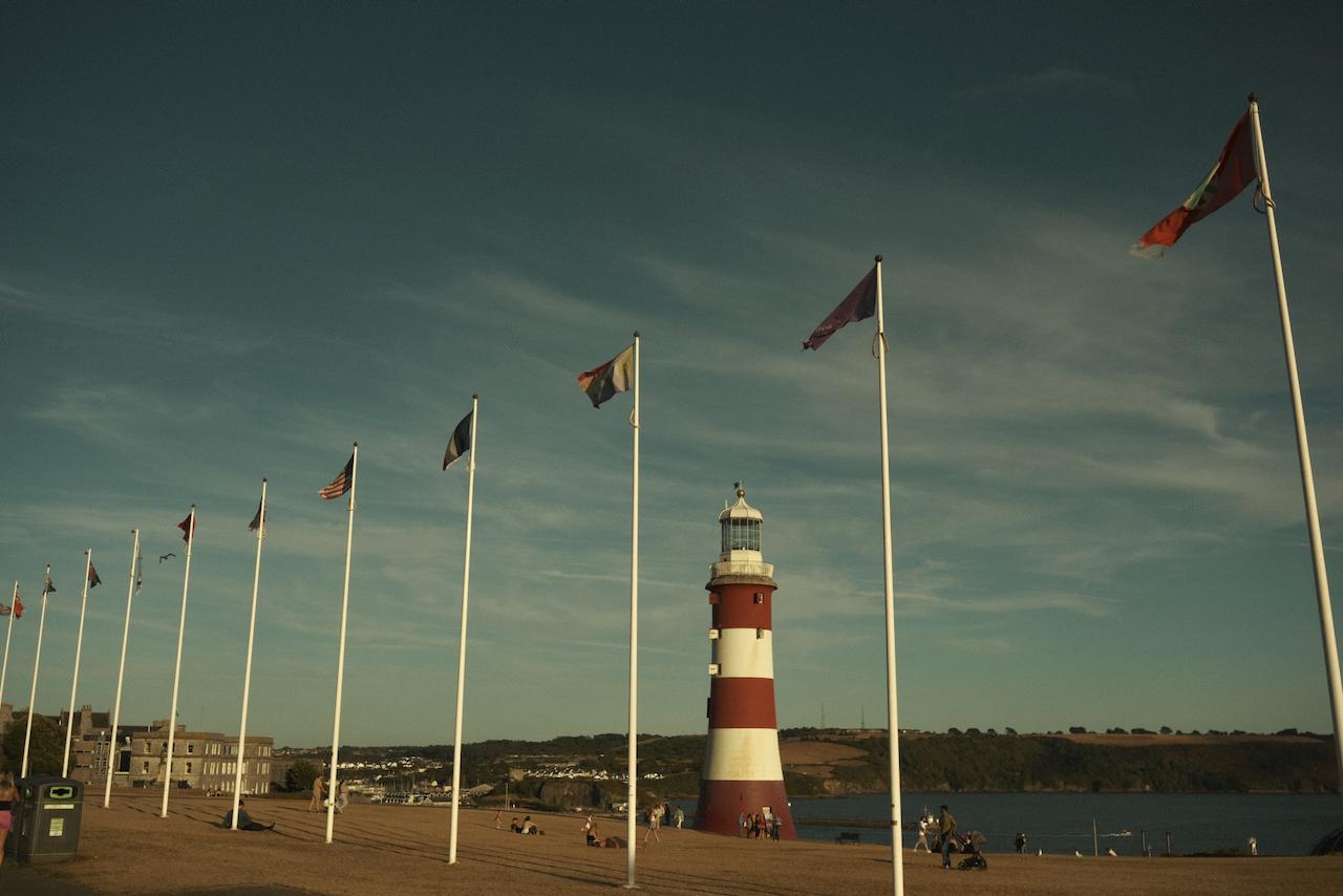 A picture of plymouth Hoe. IN the foreground are a number of flag poles with flags, in the middle ground is Smeatons Tower, and in the background is the break water.