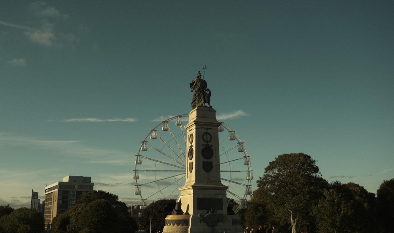 A picture of a large sculpture with a Ferris wheel behind. The sky contains wistful clouds.