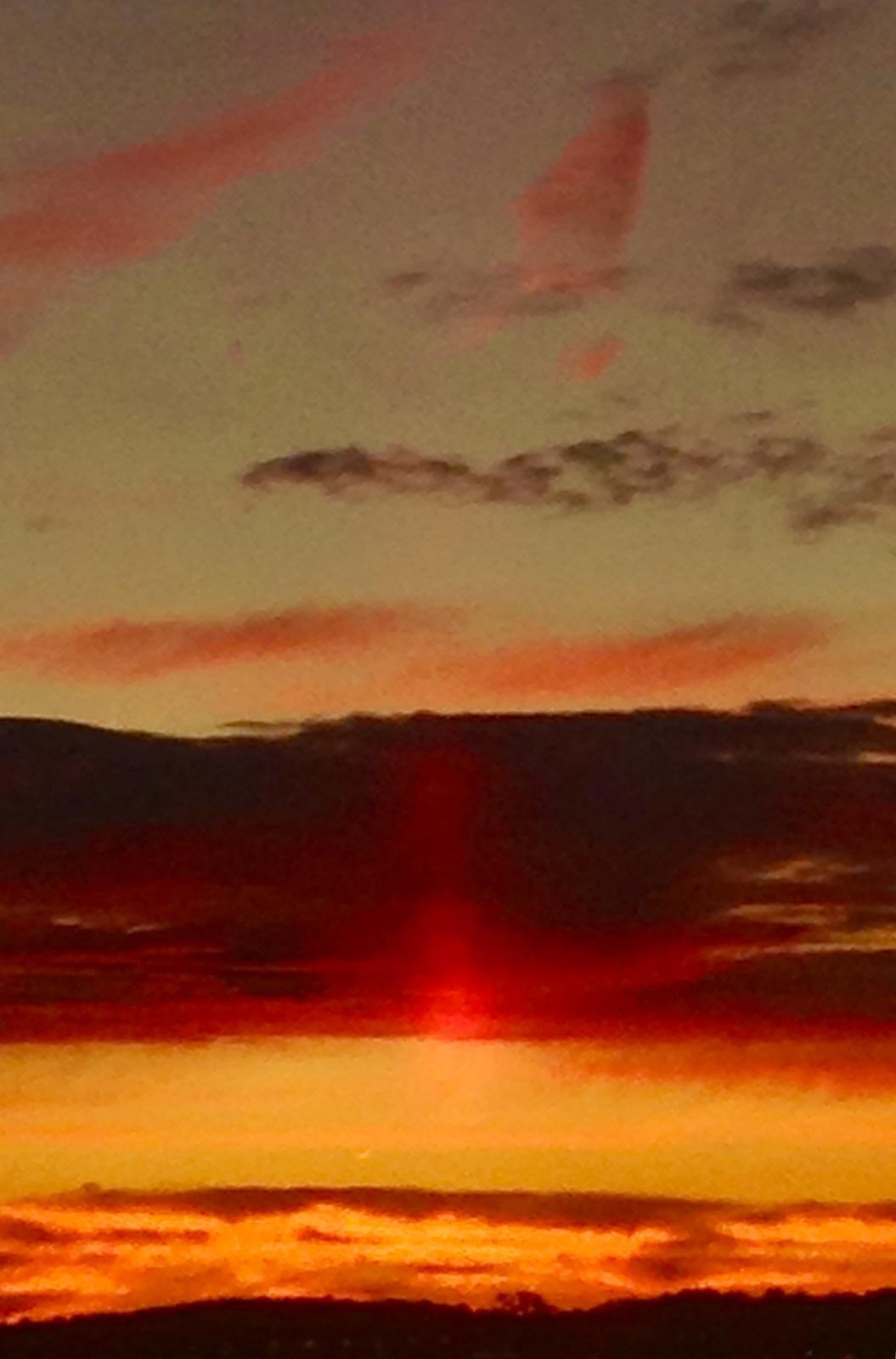 Color image of a sunset on Atwood Lake, Tuscarawas County, Ohio. USA. Image shows clouds in orange and gold and red and gray pierced by the sun toward its setting, with a pale blue sky above and a hillside covered with trees on the horizon.