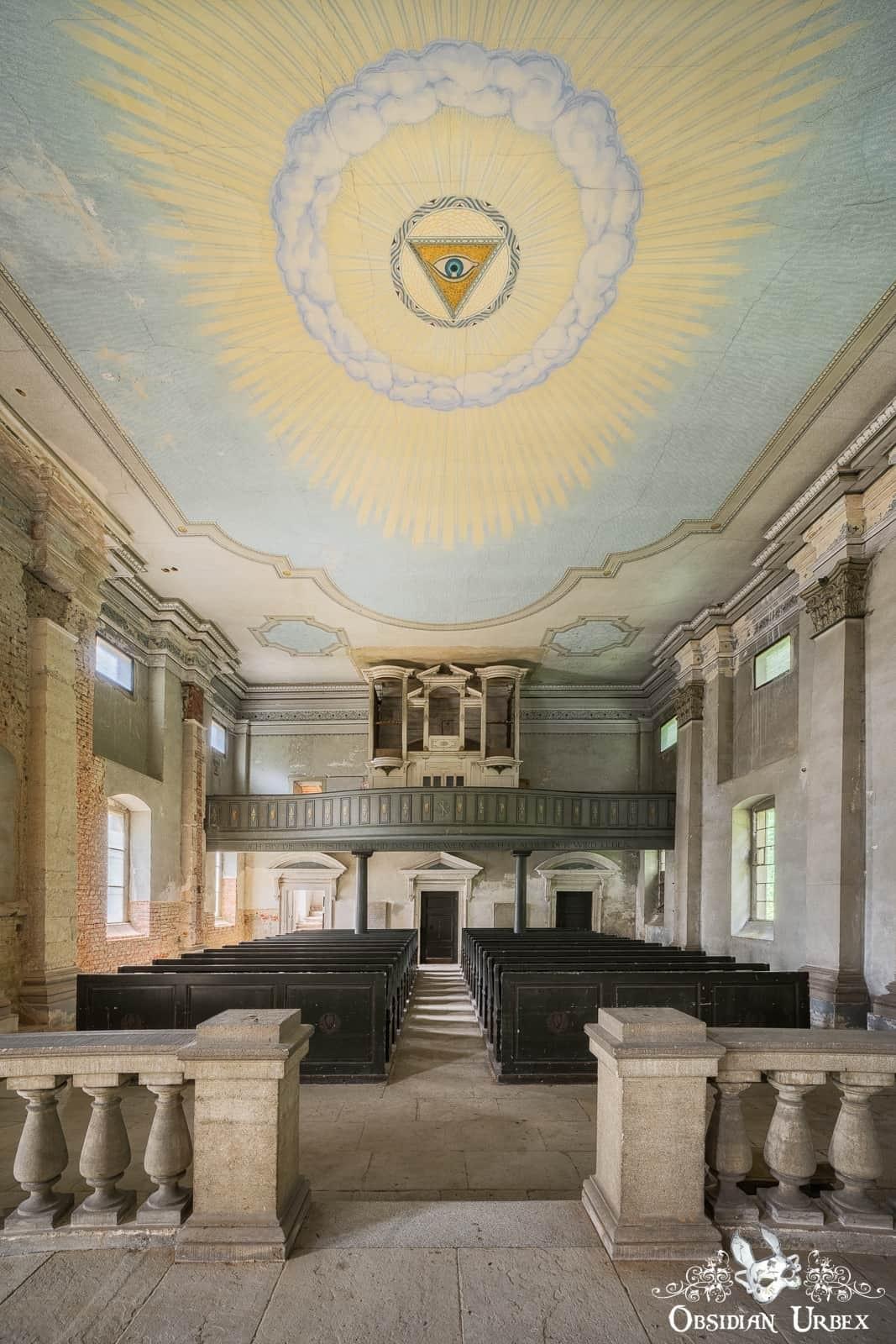 Interior of an abandoned church with an Eye of Providence ceiling mural, rows of empty pews, and an organ loft.