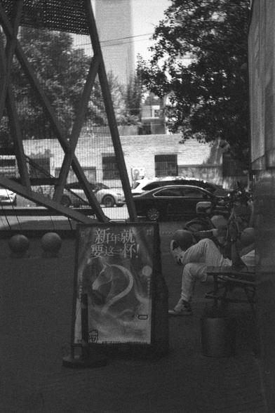 Ilford HP5 Plus 400 (FF)

English
A black and white photograph of an urban street scene. A partially seen person sits on a bench resting. An AD campaign stand is to the left of the person. In the background some high-rises can be seen. In the not so distant back, a car park is right behind a metal grid-like structure.

中文
一張城市街景的黑白照片.一個人部分可見.此人坐在椅子上休息.他的左邊有一塊廣告看板.背景中有幾棟高樓.在不遠處,有一個停車場,就在一個金屬網狀的建築後面.
