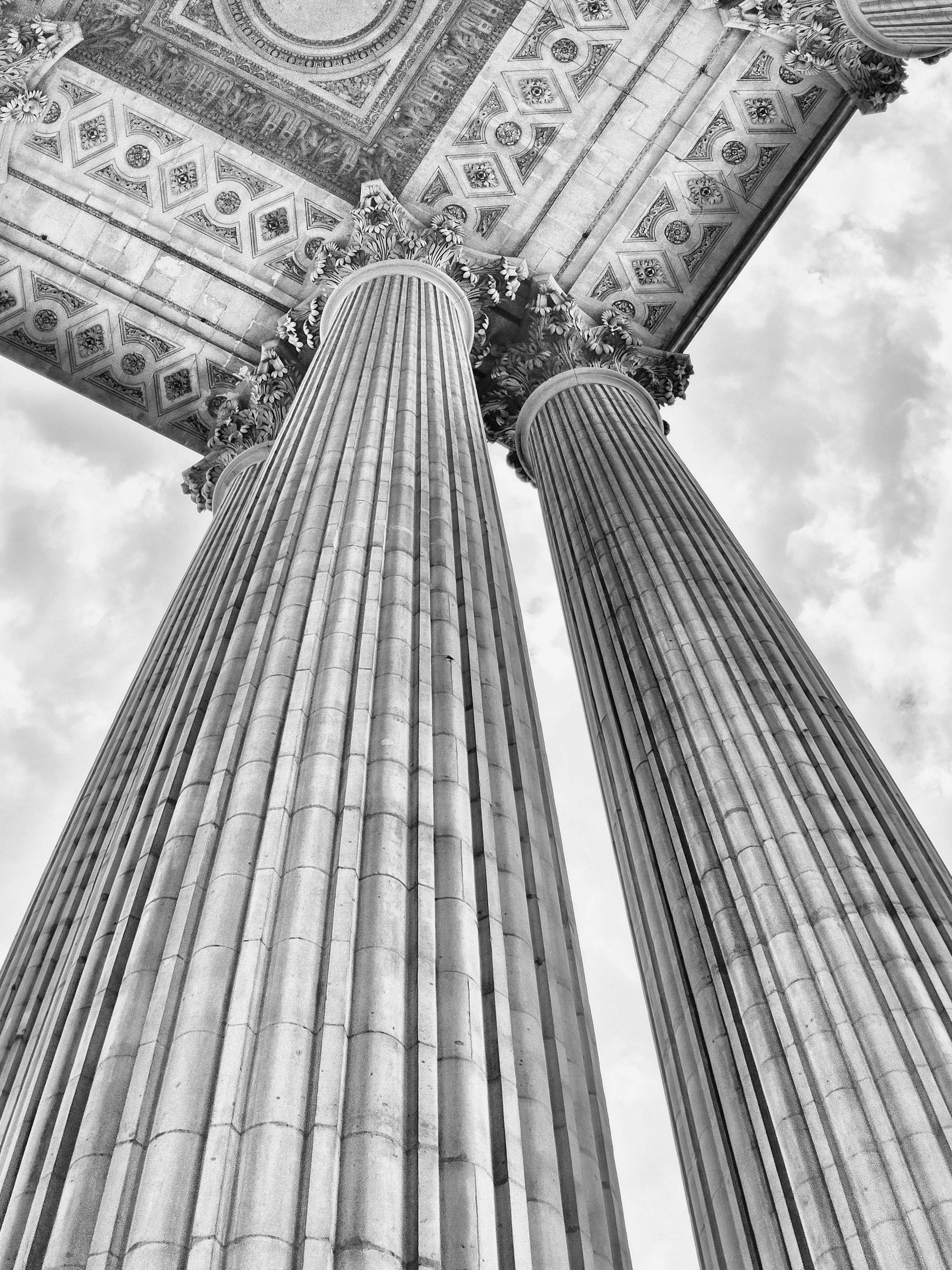 A black and white low-angle photograph that captures classical architectural columns, emphasizing their height and intricate decorative details against a vast, cloudy sky backdrop. The composition utilizes a dramatic worm's-eye view, looking straight up at the monumental columns and the ornate ceiling structure they support. The two primary columns dominate the foreground, stretching vertically through the frame to create a sense of immense scale. The perspective converges slightly towards the top, drawing the viewer's gaze from the textured stone surfaces upward toward the highly detailed, carved relief work decorating the underside of the structure, which frames the upper portion of the image.