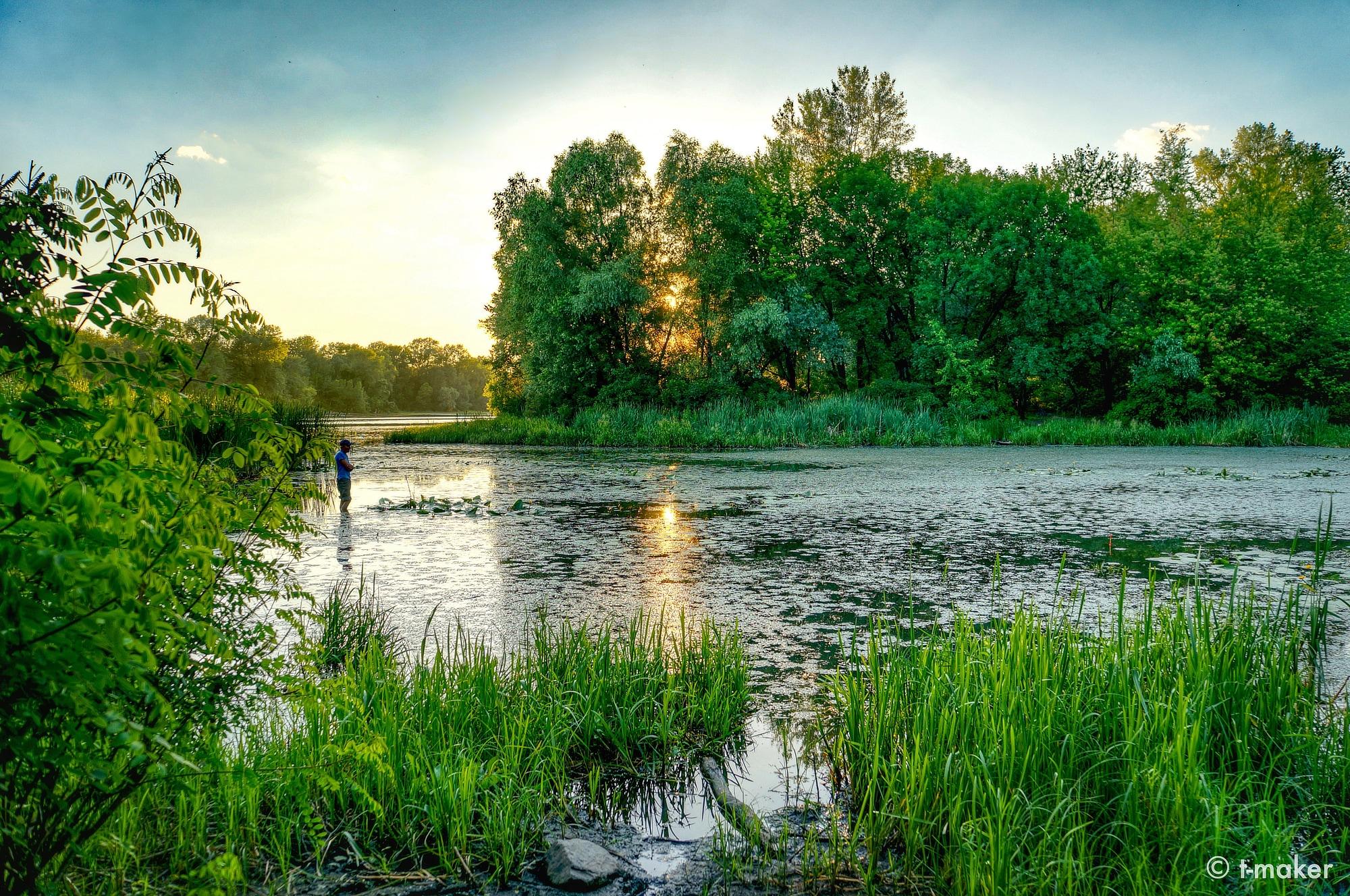 A calm river at sunset, with a fisherman standing along the bank, lush green trees and grasses, and partly cloudy sky reflected in the water.