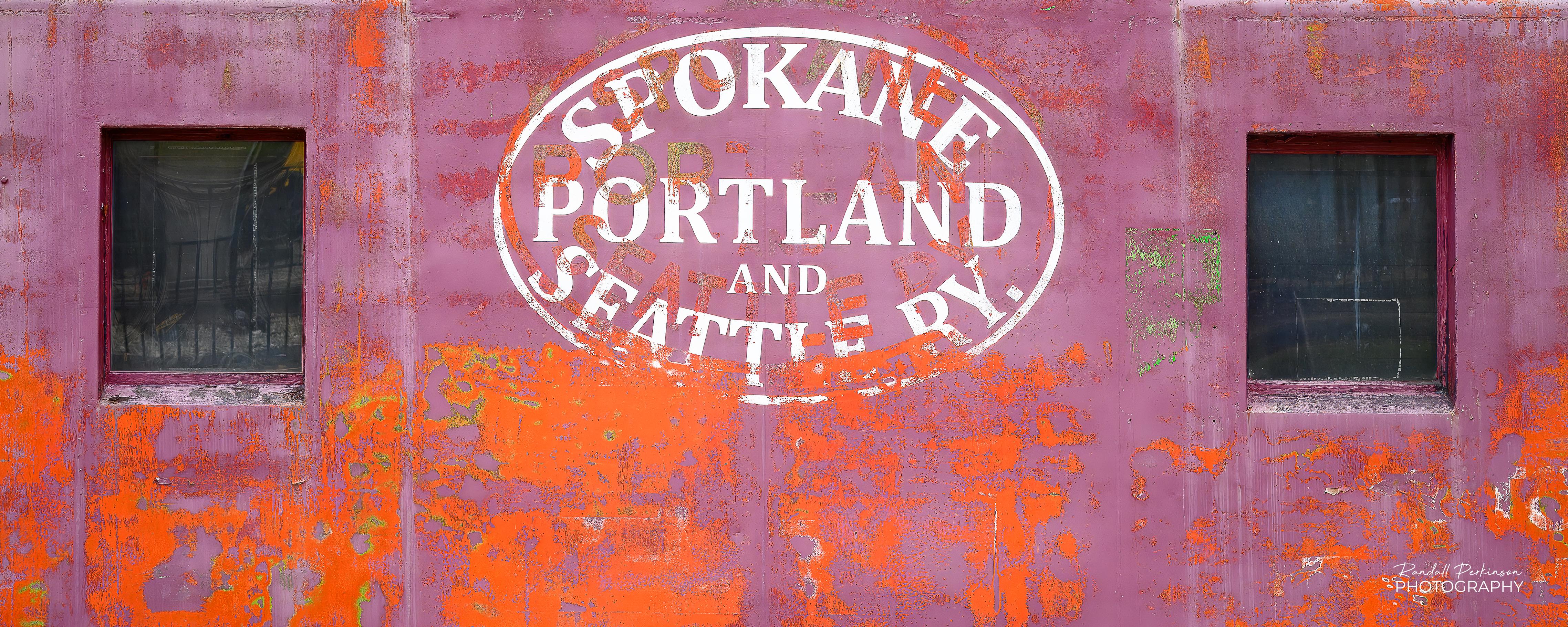 An image of the side of a rail car showing weathered, vibrant orange and purple surface feature the logo of the Spokane, Portand and Seattle Railway, know as "The Northwest's Own Railway".  The logo is framed by two windows.  The SP&S became part of Burlington Northern in 1970.