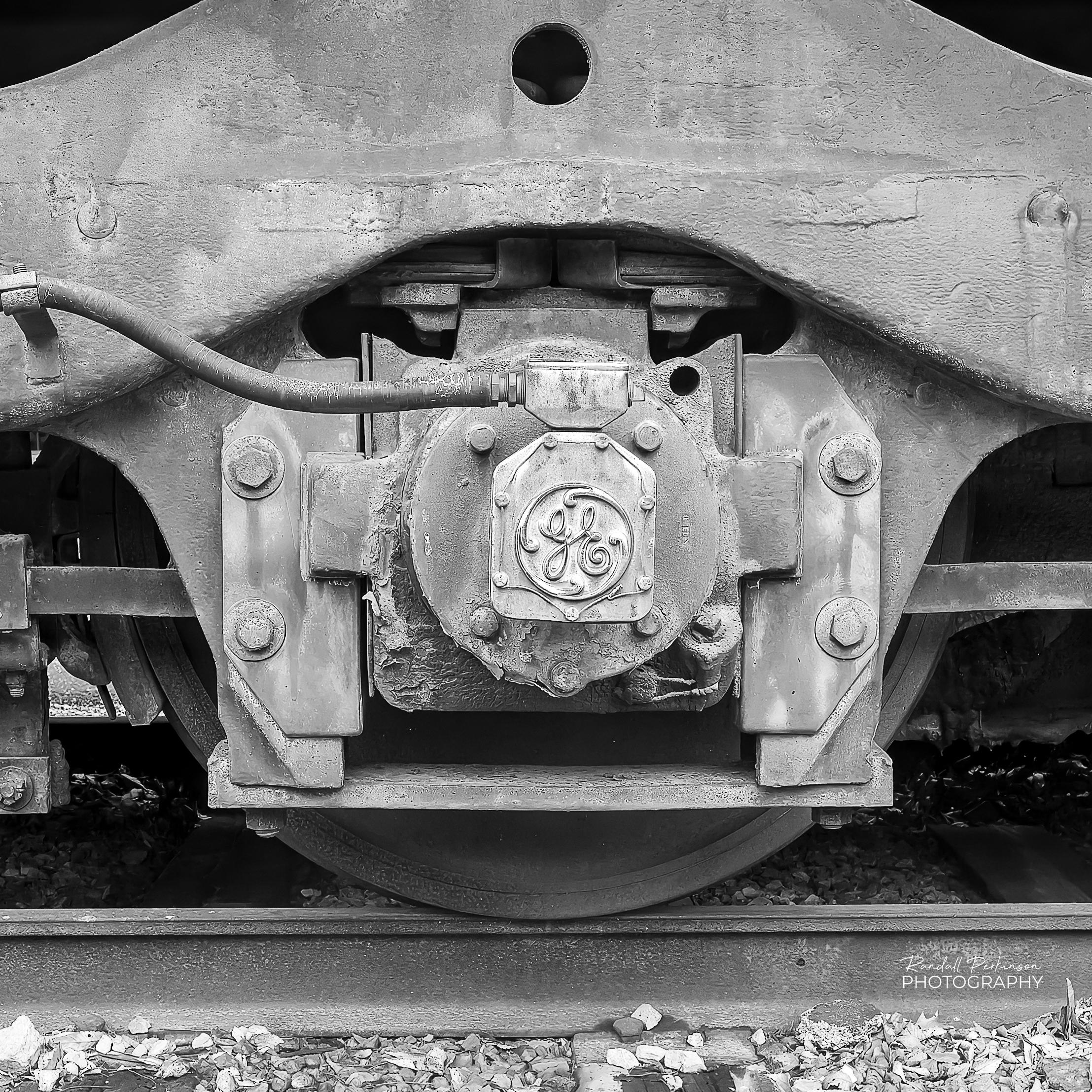 A closeup of a journal bearing/axle box assembly mounted on the wheelset of a locomotive truck.  The central cap prominently features the vintage General Electric (GE) monogram logo.