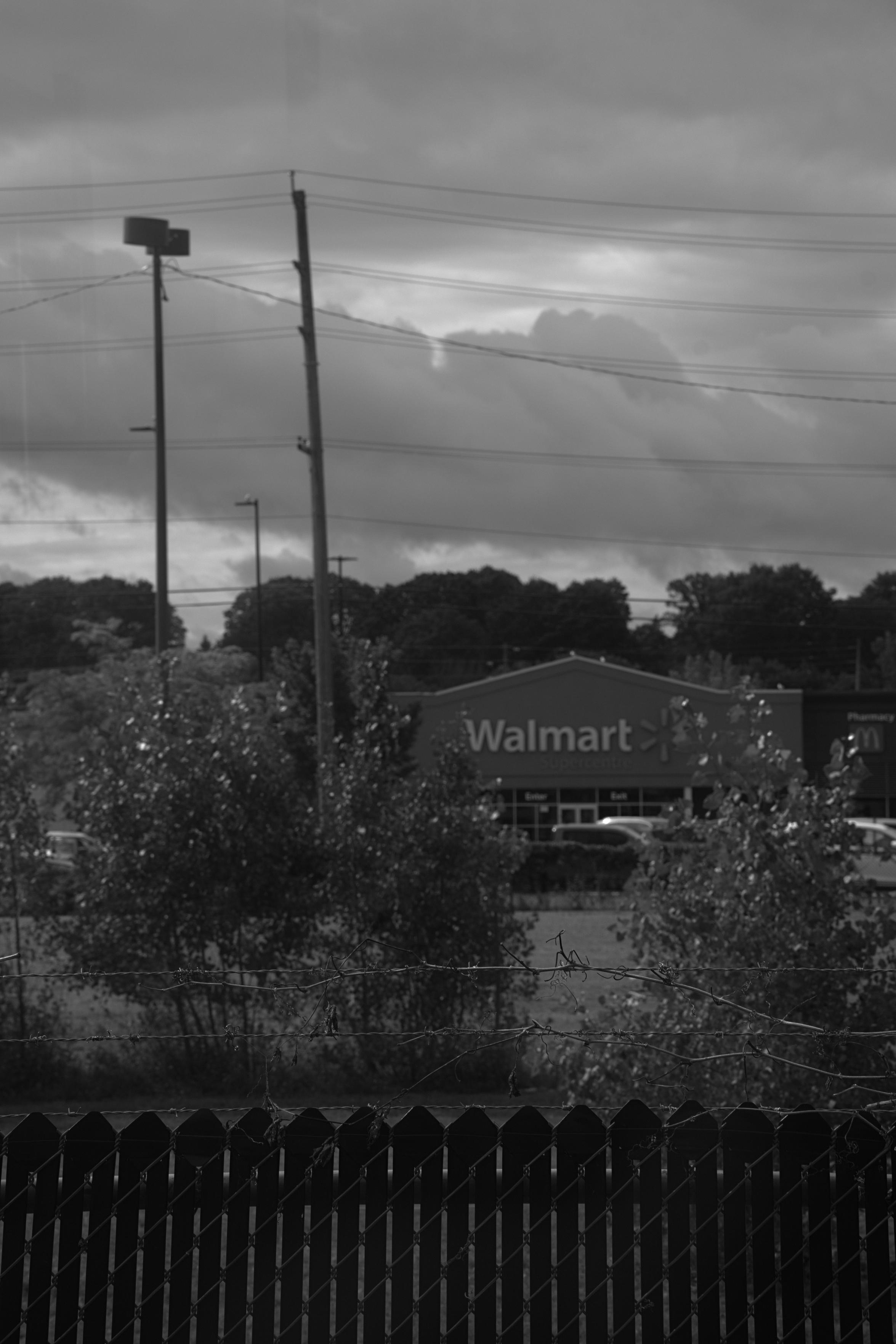 A dark fence topped with barbed wire, and a Walmart and cloudy skies in the background.