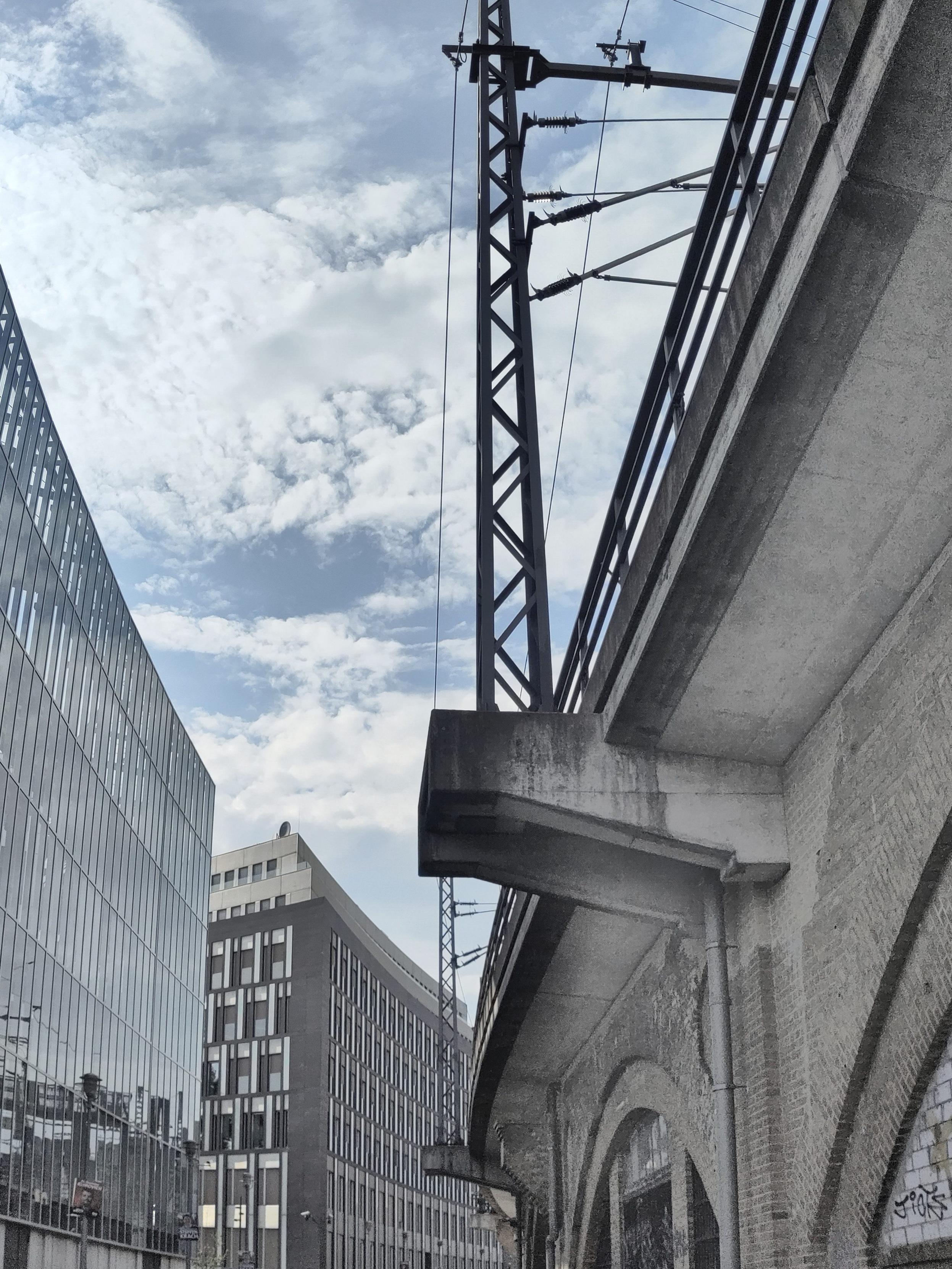 An upward view along an elevated concrete railway viaduct, seen from below at a steep angle so it recedes diagonally into the distance on the right. The viaduct has a projecting concrete ledge and rows of arched openings at street level. A tall steel lattice mast with overhead lines and insulators rises above it. On the left stand modern multi-storey buildings, including a glass-fronted facade and a brick-and-window office block. The sky is blue with scattered white clouds. The palette is largely grey with cooler tones.