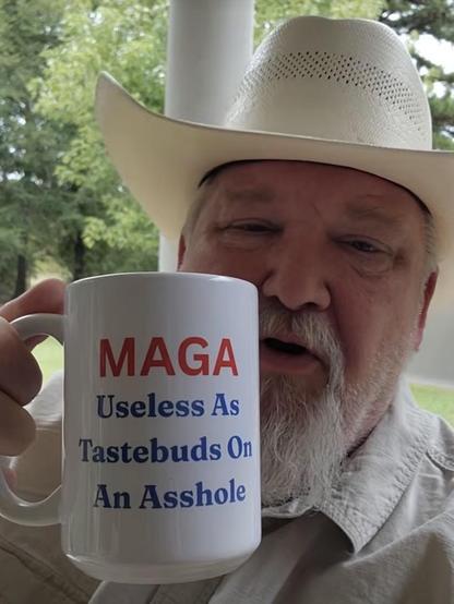 Photo of a bearded old white man in a Stetson hat holding up a coffee mug with the words, “MAGA useless as tastebuds on an asshole.”