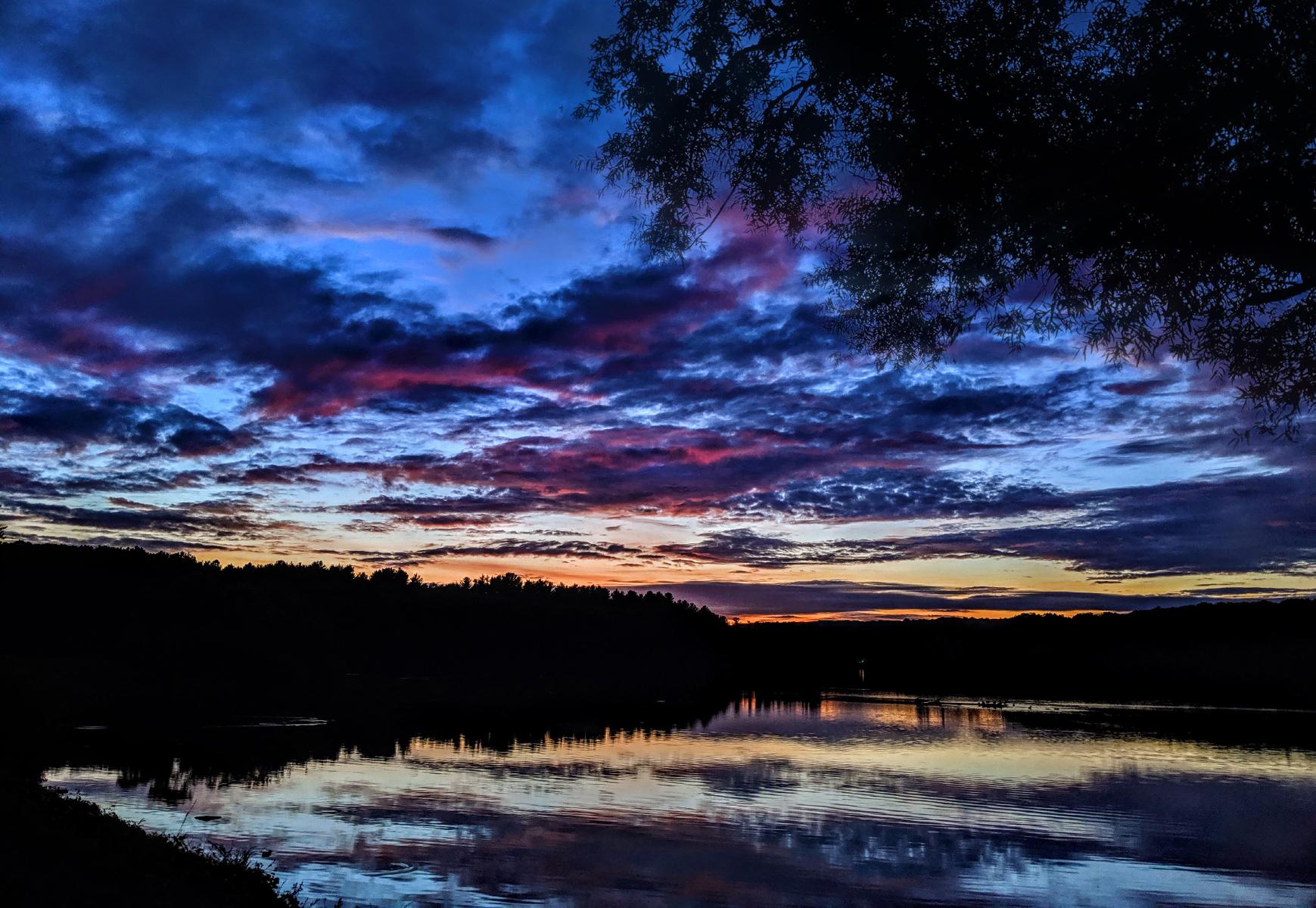Walking a footpath beside a lake in the twilight. A tree overhangs the path to our right. The far shore is wooded. The sky is a deep blue, with darker navy blue clouds. Many of the clouds are streaked with pink from the recent sunset. The horizon is a pale orange. The sky is mirrored on the gently rippled lake below.