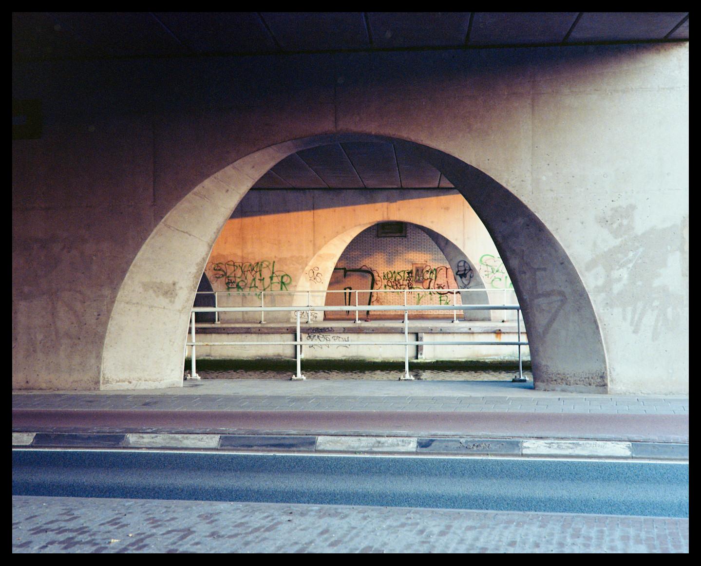A viaduct shot during early morning sun. Concrete arches, creating a bit of a tunnel effect partially lit by warm sunlight. Graffiti on the walls, with a road and sidewalk in the foreground. An obvious urban scene.

Camera: Nikon F801s
Lens: Nikkor AF 50mm f/1.8D
Film: Kodak Ektar 100