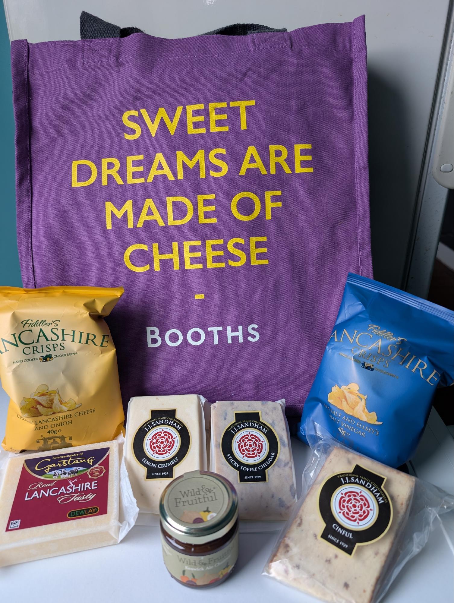A purple cloth bag and some goodies are arranged on a white surface. The bag reads "Sweet Dreams Are Made Of Cheese" in yellow above the name Booths in white. The goodies consist of two packets of crisps - Cheese and Onion, and Salt and Vinegar - plus a small jar of chutney and four sealed cheeses, all from Lancashire. One cheese is a standard block of white, while the other three are wedges, decorated with the red rose of the county, and with interesting flavours: Cinful (no idea), Lemon Crumbly, and Sticky Toffee Cheddar.