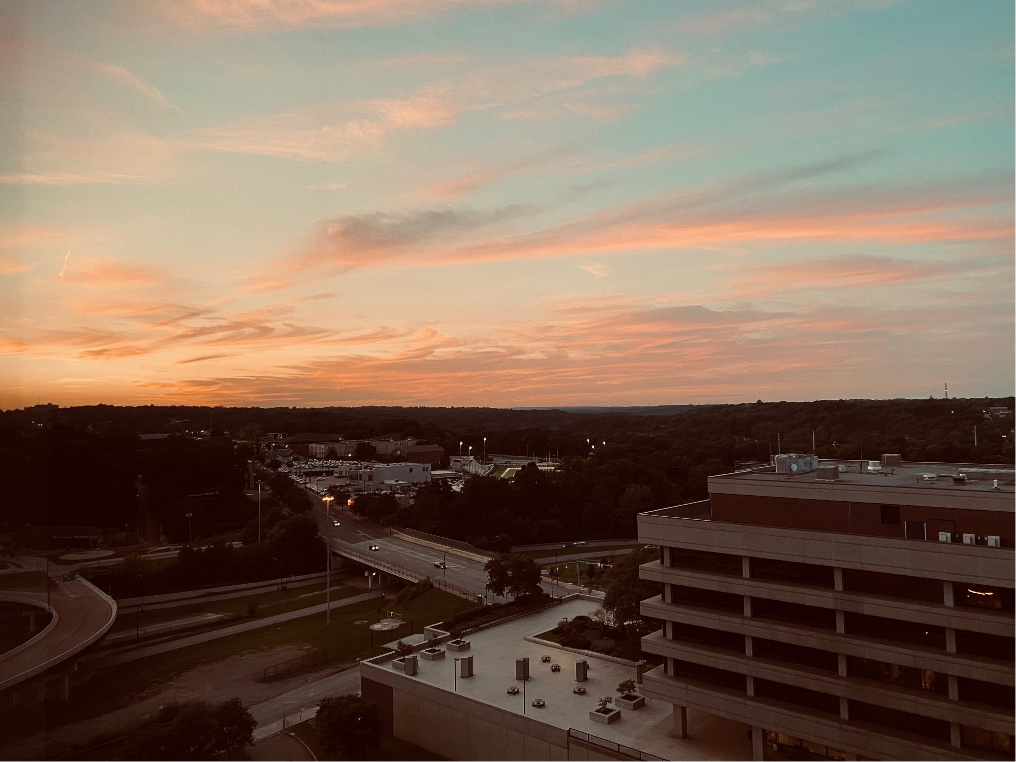 Color image of twilight, the sun just having set in Akron, Summit County, Ohio, USA. Image shows light clouds underlit by the set sun, aglow in oranges and yellows, blue sky above, and the building of the city below the horizon cast into the darkness of dusk.