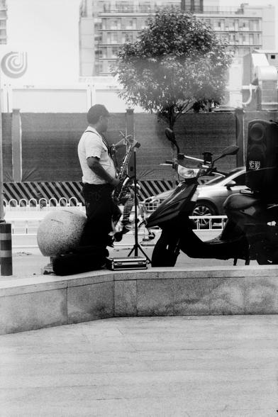 ERA 100 (FF)

English
A black and white photograph of a street scene. A person is playing a saxophone by the side of the street. Across the street there is a building behind a tree. The person stands by a motorcycle.

中文
一张街景的黑白照片.照片中一个人在演奏萨克斯风.演奏者站在一辆电瓶车遍.街对面有一栋楼房,前面是一棵树