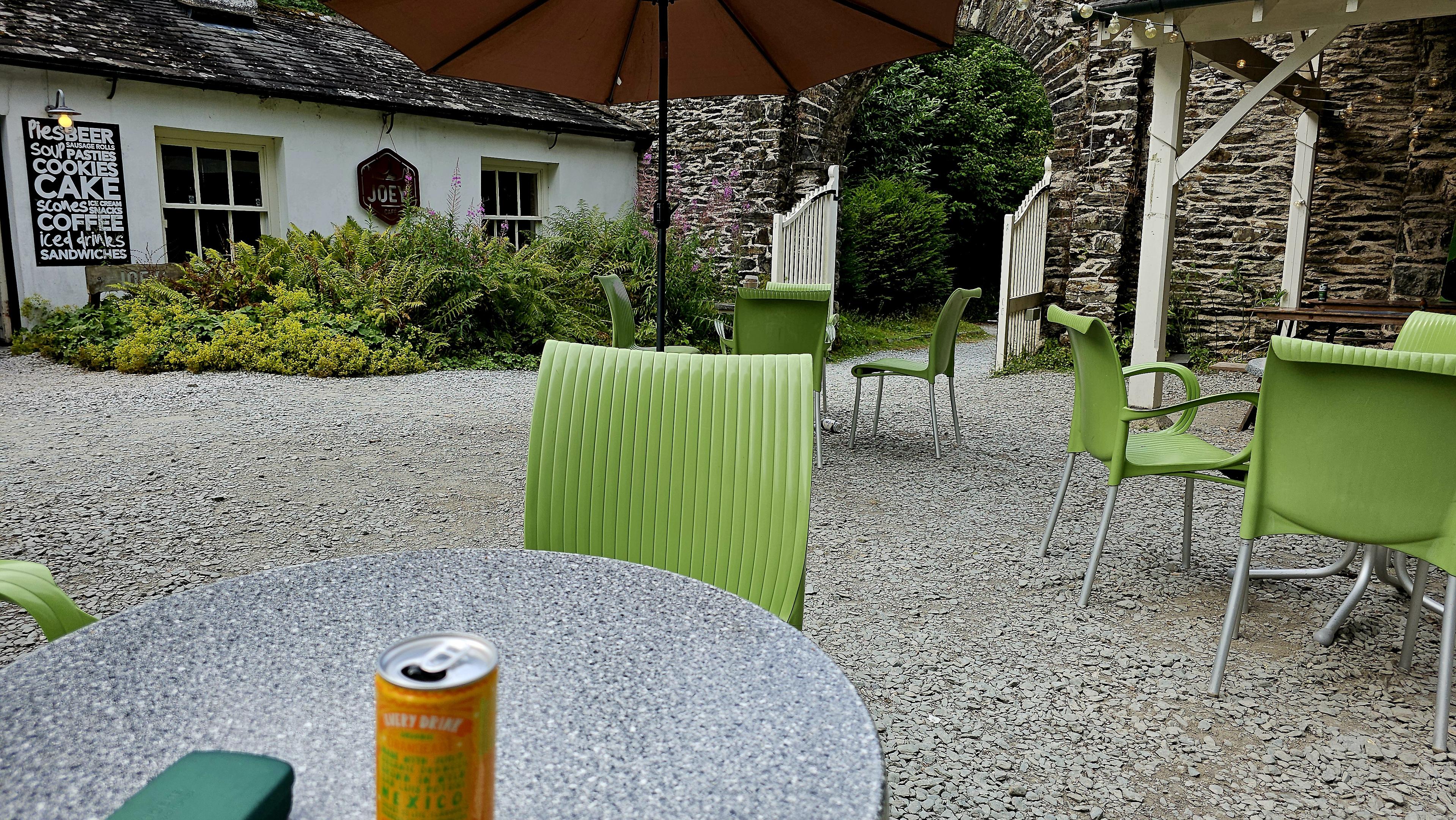 Outdoor cafe tables, with green, plastic chairs and an umbrella, stand on grey gravel. A low building, with two windows and handwritten signs, stands to the left. A high, stone wall, with white, wooden gates, stands to the right.