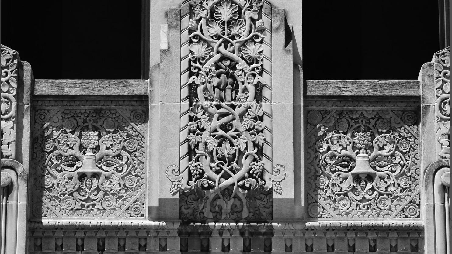 Color image of design elements in the entablature surmounting the grand front entrance into the 1930 Ohio Bell Telephone Building, an Art Deco era building in Akron, Summit County, Ohio, USA. Image shows the intricate stone carving work featuring a center panel filled with grapevines surrounding a cantlestick telephone, and with the side panels continuing the grapevine motif, each surrounding bells, the corporate symbol of the Bell Telephone Company super corporation.