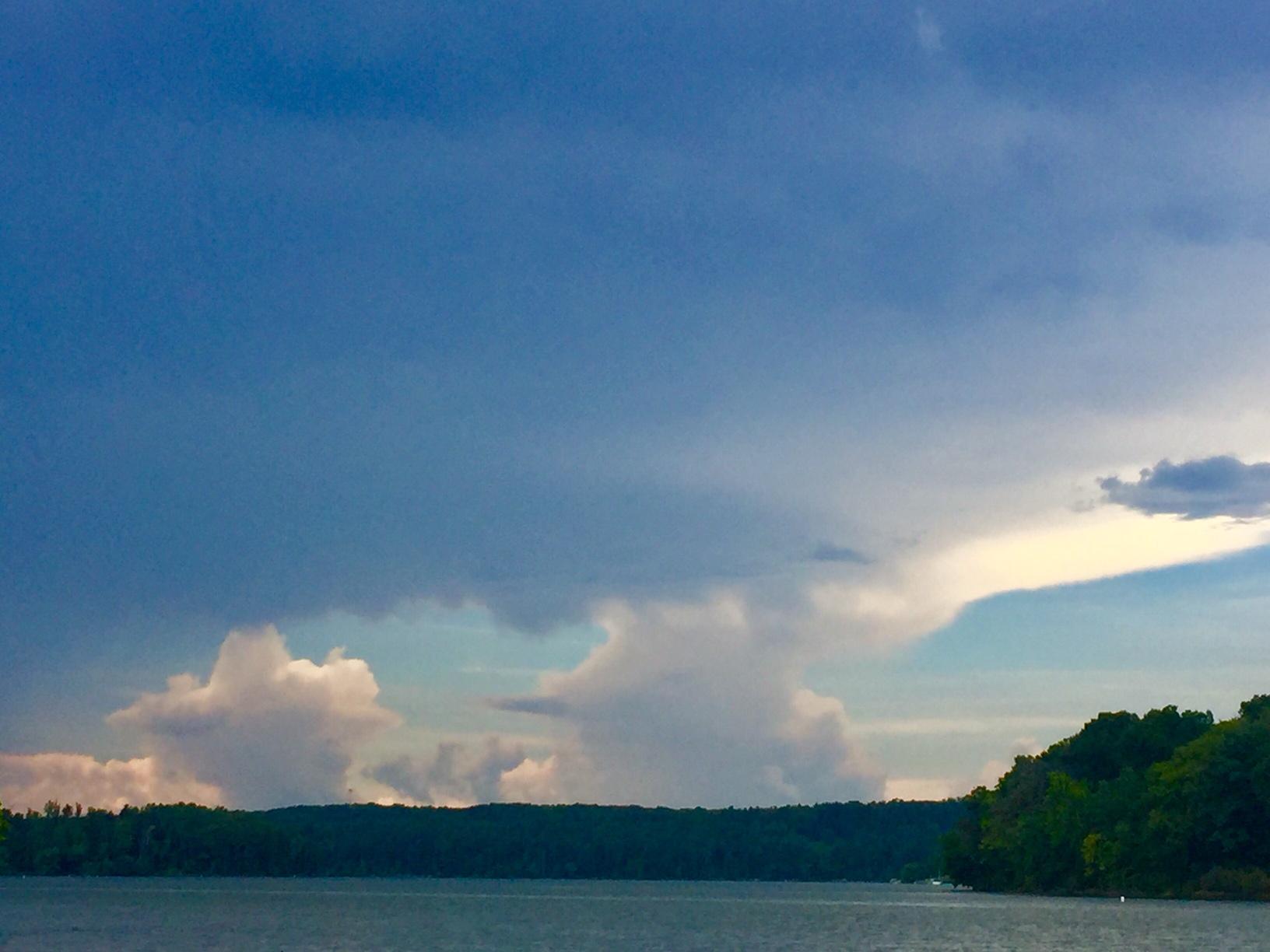 Color image, sunset, August, Atwood Lake, Carroll County, Ohio, USA. Image shows cumulus clouds on the western horizon as the sun sets over the lake, blue sky above, ripples on the lake below, trees hugging the banks and hillsides.
