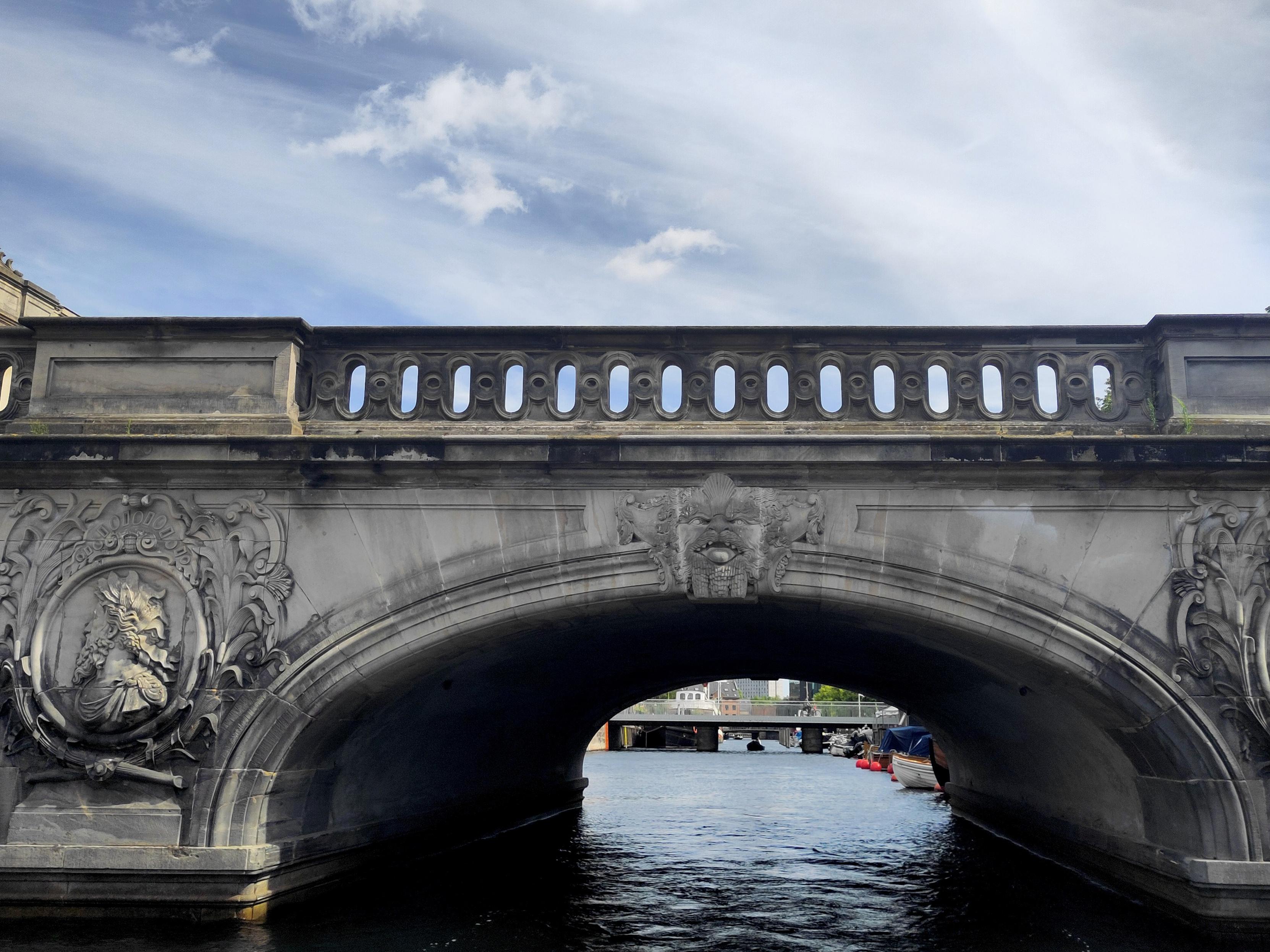 An ornate old stone bridge arching over a canal, viewed from water level. The dark grey stonework is richly decorated with Baroque carvings, including foliage scrolls, oval medallions and a grimacing mask face at the top of the arch. The parapet has a pierced balustrade with oval openings. Through the arch, the canal continues to a second, flatter bridge in the distance, with moored boats and buildings along the banks. The sky above is blue with white clouds.