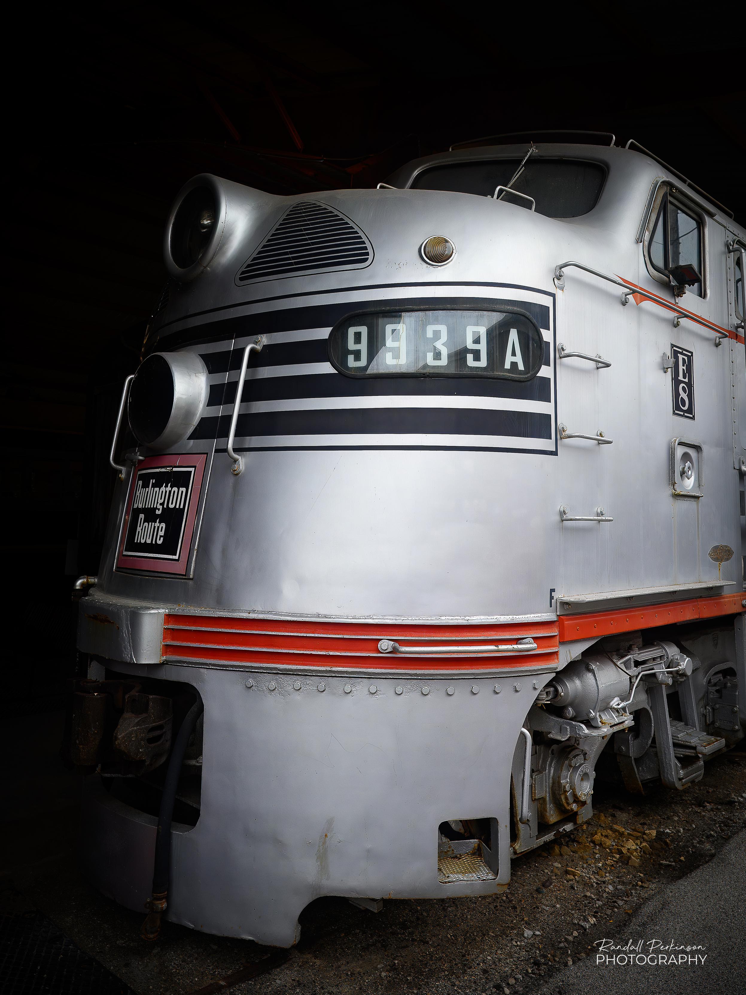 Close-up three-quarter view of a vintage silver and red Burlington Route diesel locomotive, number 9939A.  The train features a classic streamlined nose, horizontal black and red stripes on front.  It is on display against a dark background.