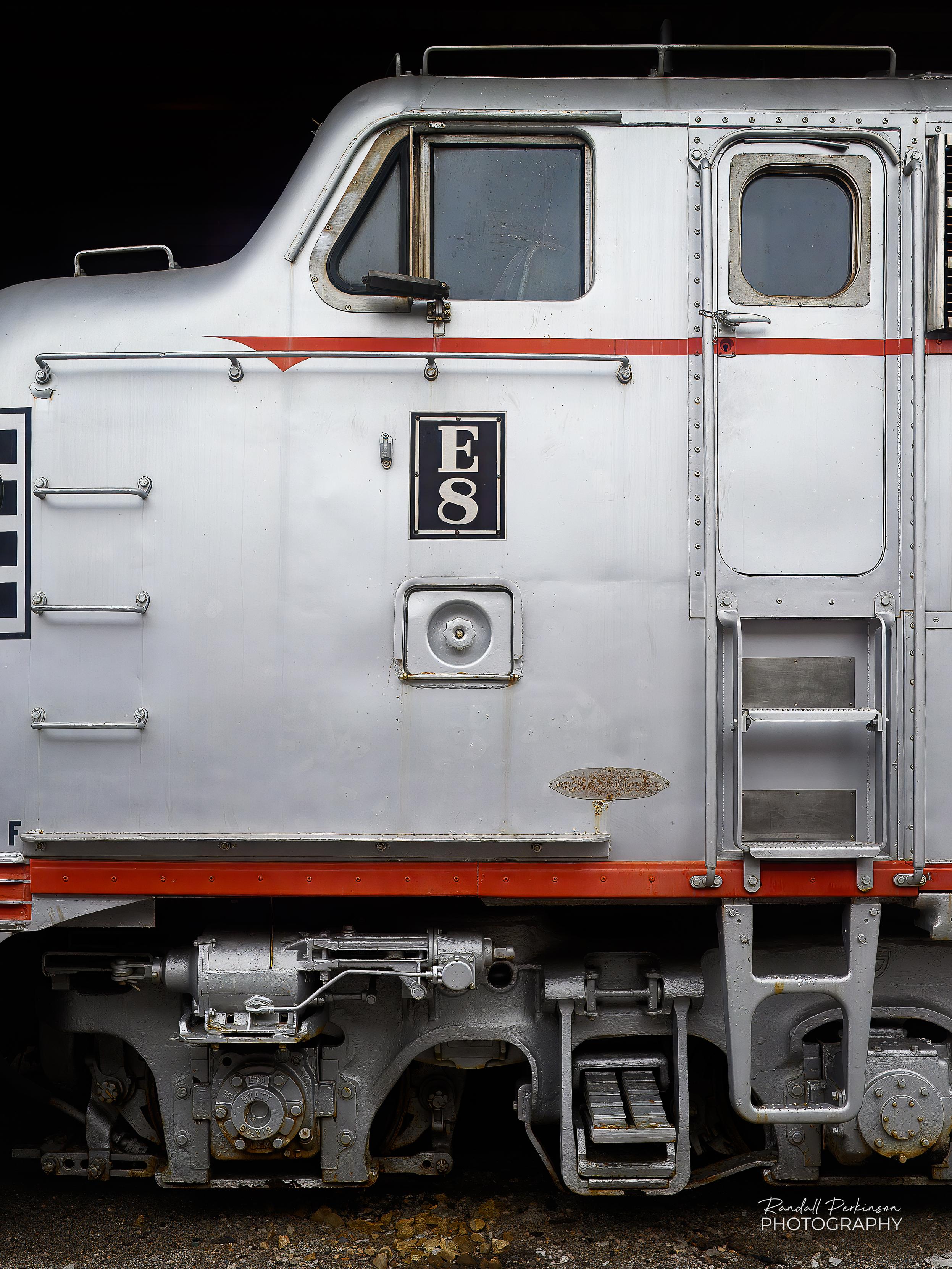 Close-up side view of a vintage silver and red Burlington Route diesel locomotive, number 9939A. The train features a classic streamlined nose and horizontal red stripes. It is on display against a dark background.