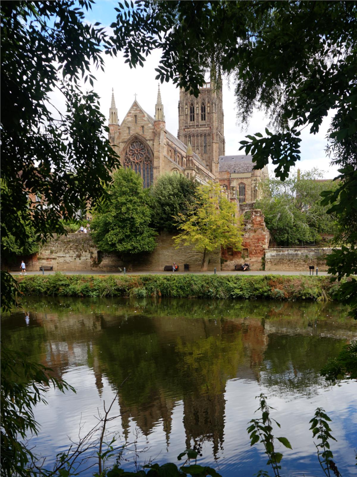Worcester Cathedral viewed across the River Severn through the trees.
The Cathedral is reflected in the calm waters of the river. The sky behind mainly cloudy but with a patch ob blue at the top