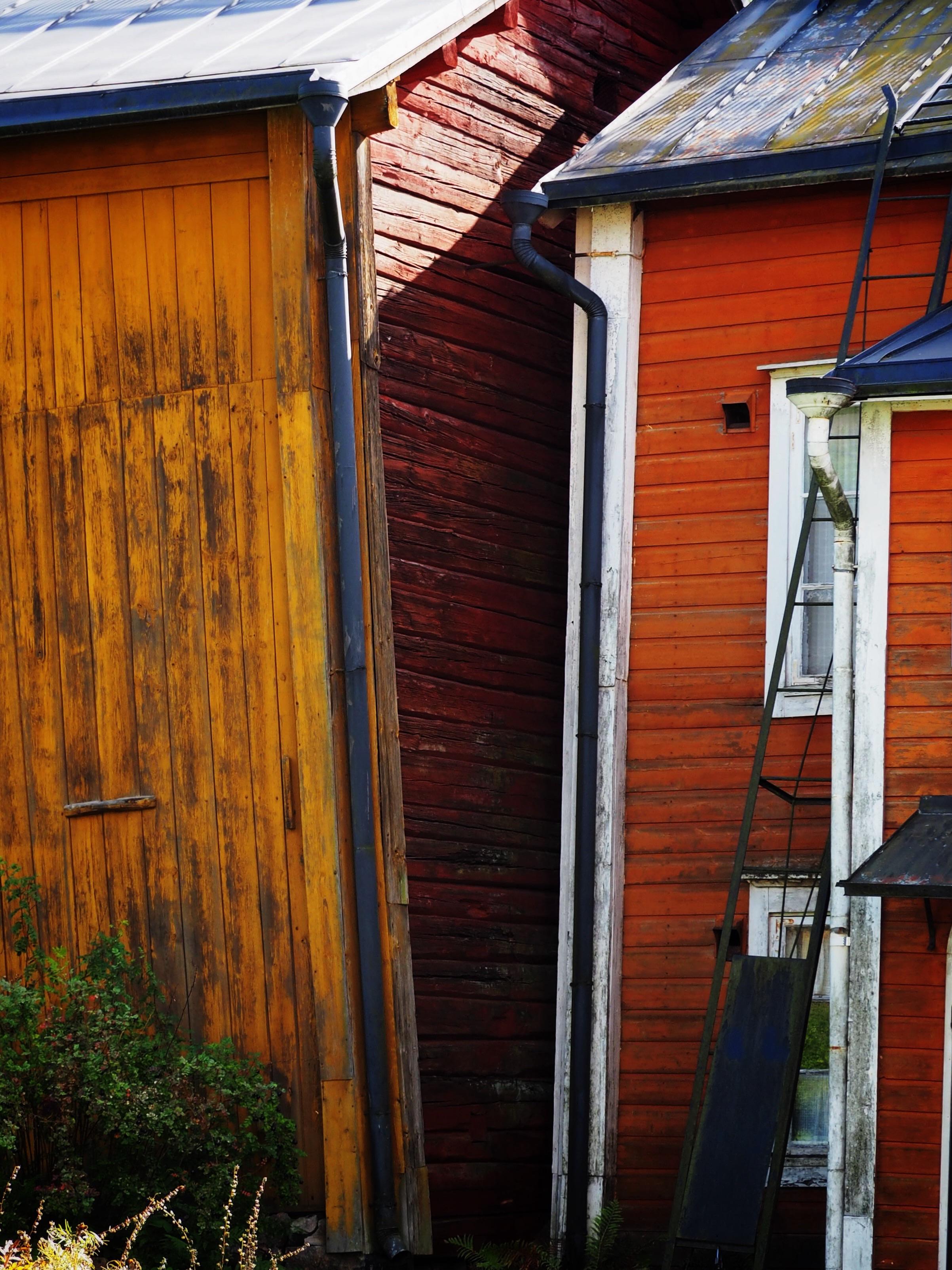 A close-up of two wooden buildings with colorful exteriors: one side is painted yellow and the other red. They are positioned closely together, with visible textures of the wood and a ladder leaning against the red building. Green foliage is present in the foreground.