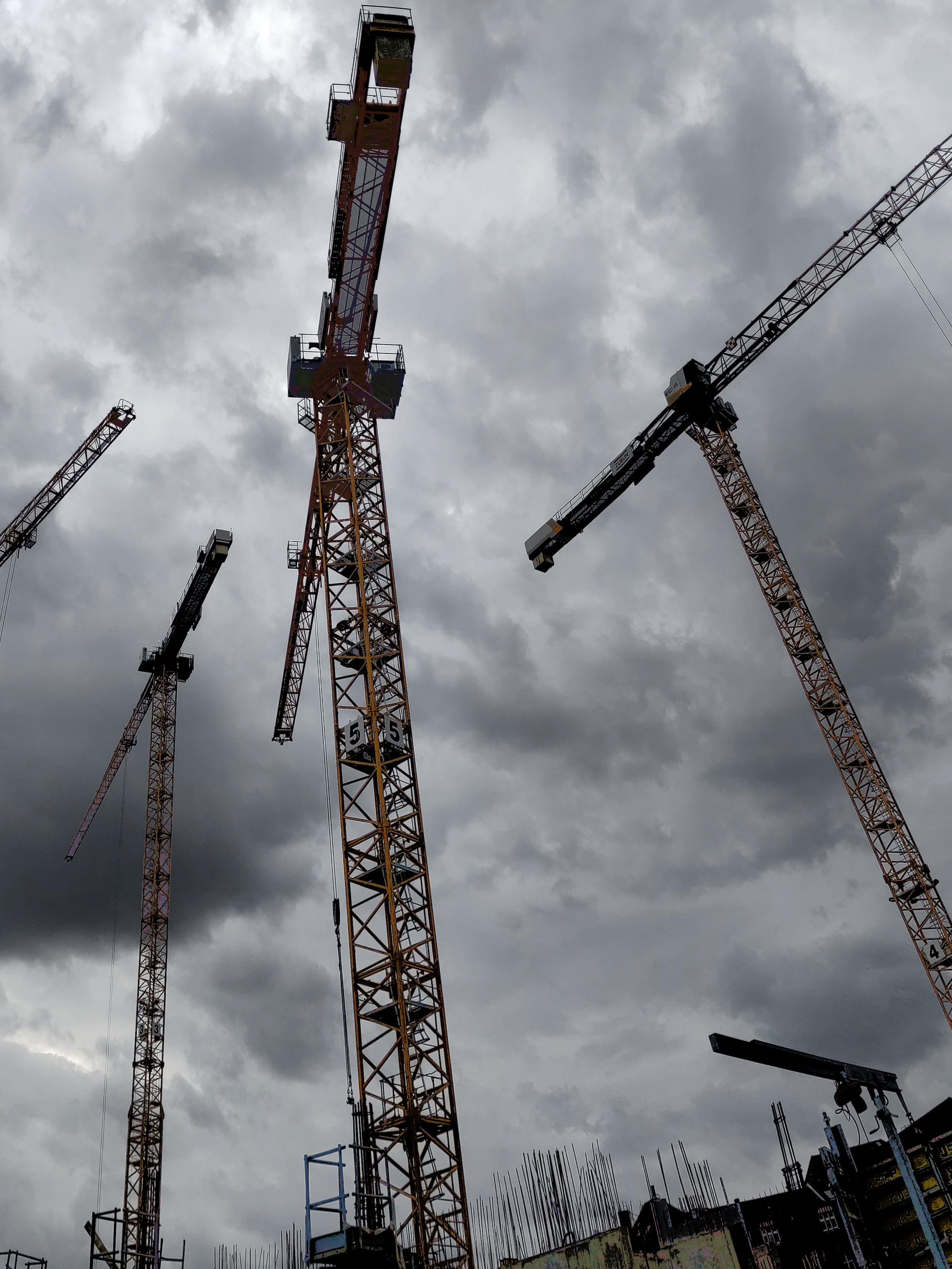An upward view of several tall tower cranes on a construction site against a heavy, overcast grey sky. Four lattice cranes with horizontal jibs rise at different angles across the frame, their steel towers appearing dark against the cloud. At the bottom, protruding reinforcement bars, scaffolding and the tops of buildings under construction are visible. The mood is dark and monochromatic with muted brown and grey tones.