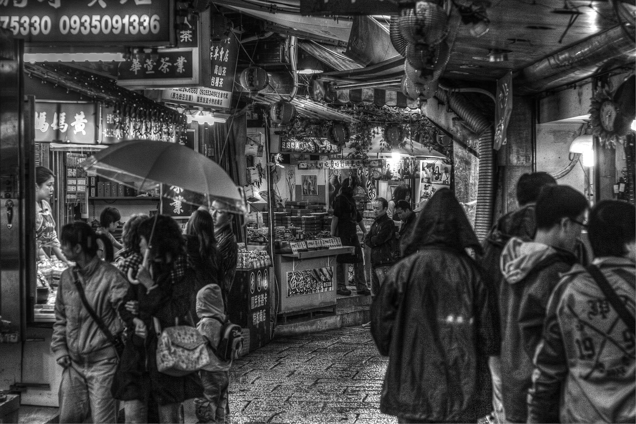 It's a dark, black and white scene, with high contrast showing off deep dark shadows and bright points of lighting. We're in a narrow alley lined with small shops with open fronts and stalls on display, some jutting out slightly into the alley space, all with an awning of some kind above as the weather warrants it. The alley's narrow enough that the overhead coverings from the small shops either side almost form a complete barrier to the elements but we can tell it's raining from the dampness on the ground and the done-up coats and raised umbrellas present. The alley heads forward from us only a half dozen metres past a couple of premises either side before it curves around to the right and is lost behind the shops there and the groups of people who are walking along each side, perusing what's on display, and opening up a narrow space to pass between them all. The signs have Chinese writing, some of them illuminated, and there are round, Chinese lanterns strung up along the front of al