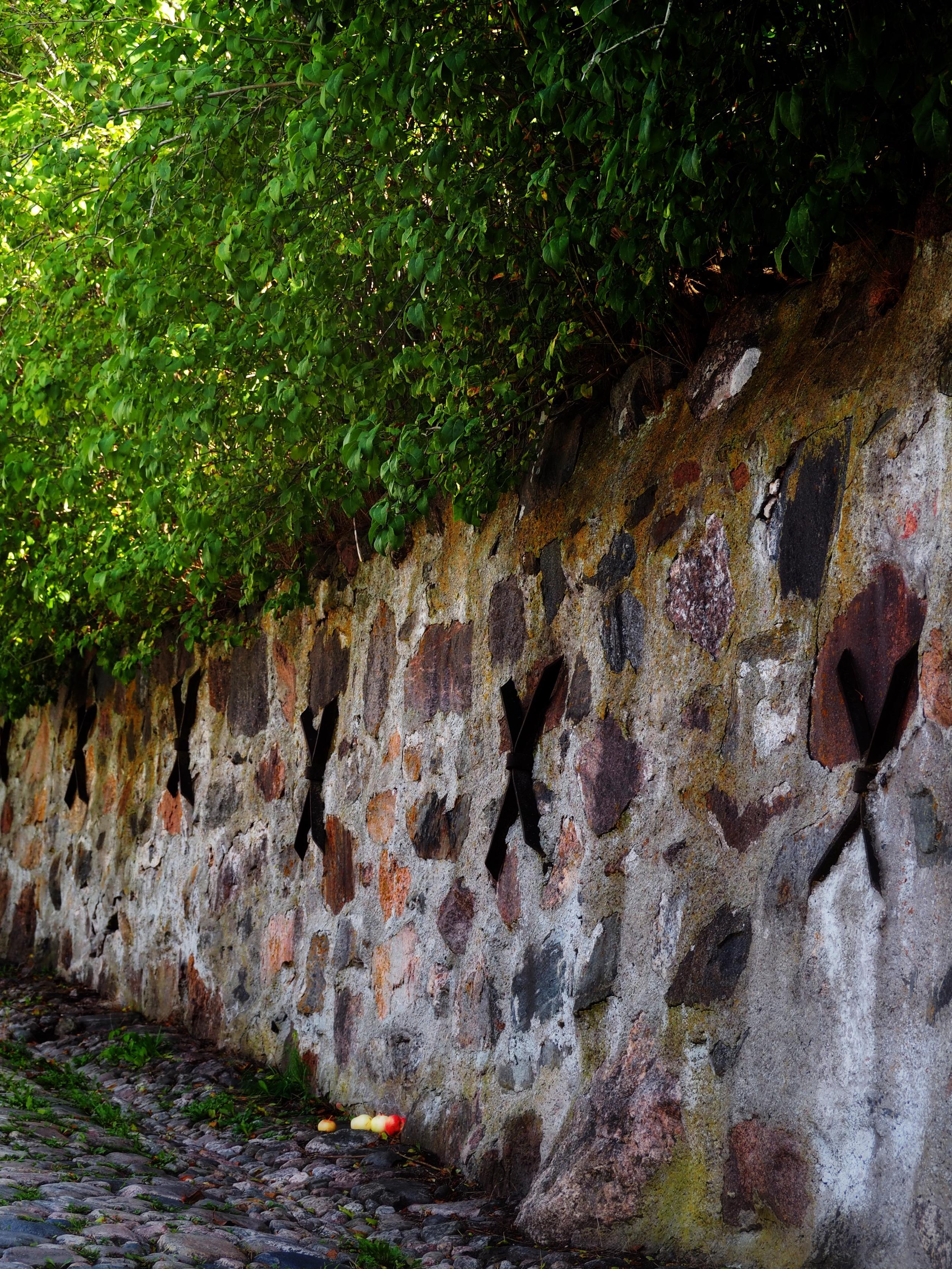 A stone wall partially covered with greenery features several black X-shaped steels on its surface. In front of the wall, there are a few apples on the ground beside a cobblestone path.