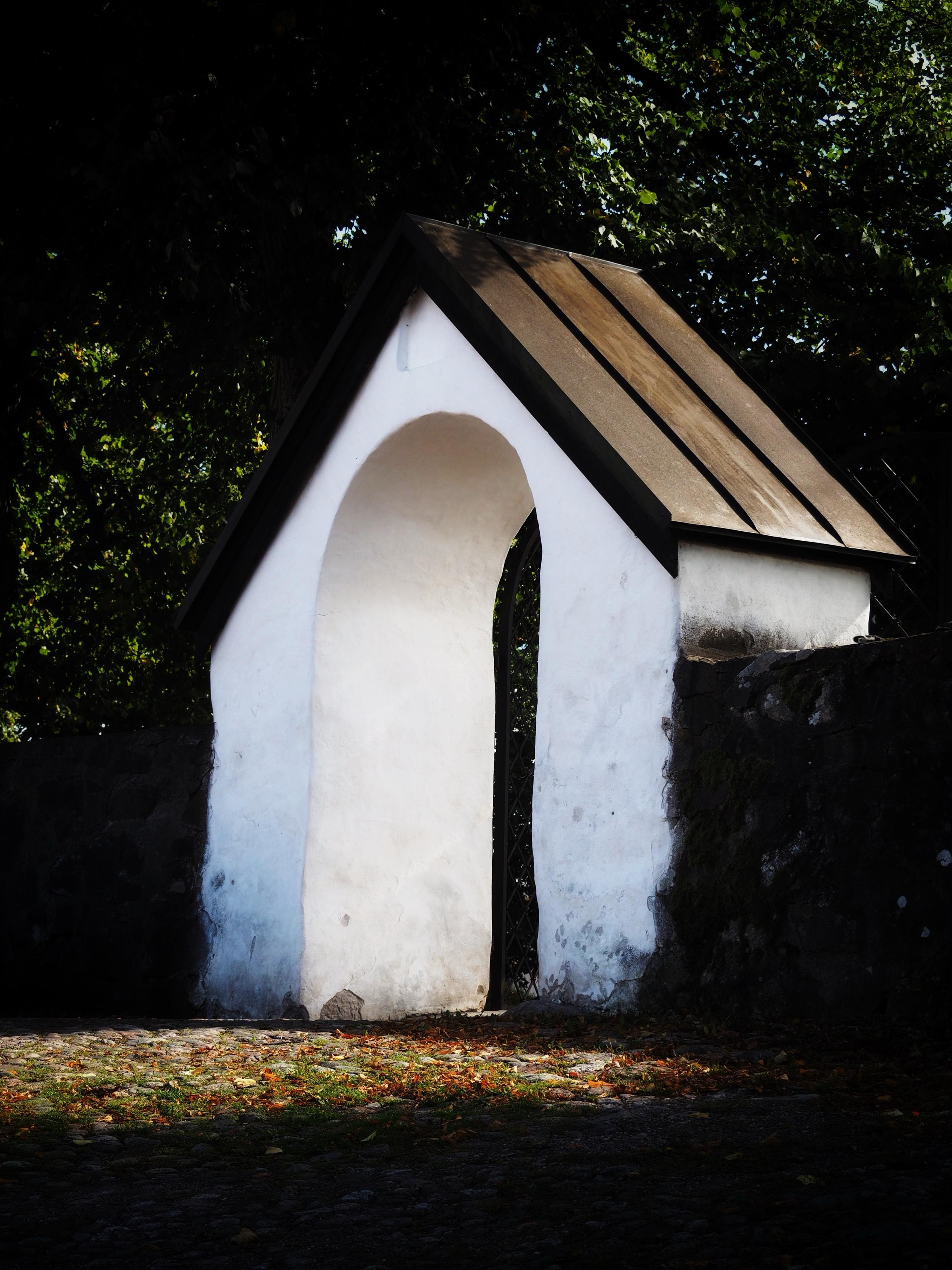 A white, arched gateway with a sloped roof, set against a backdrop of trees and a cobblestone path, with some fallen leaves scattered on the ground.
