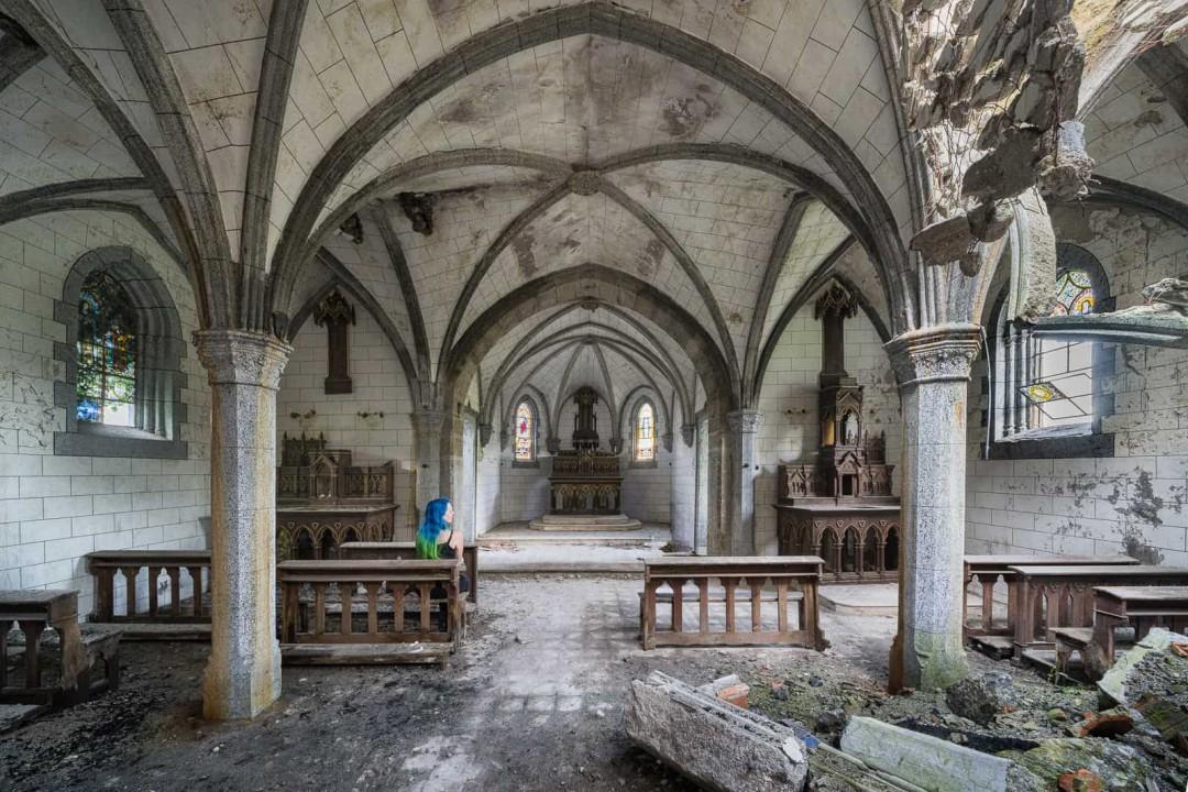 An interior shot of an abandoned stone chapel, featuring vaulted ceilings and scattered debris. A girl with blue and green hair sits to the left on the pews, looking across the vacant space